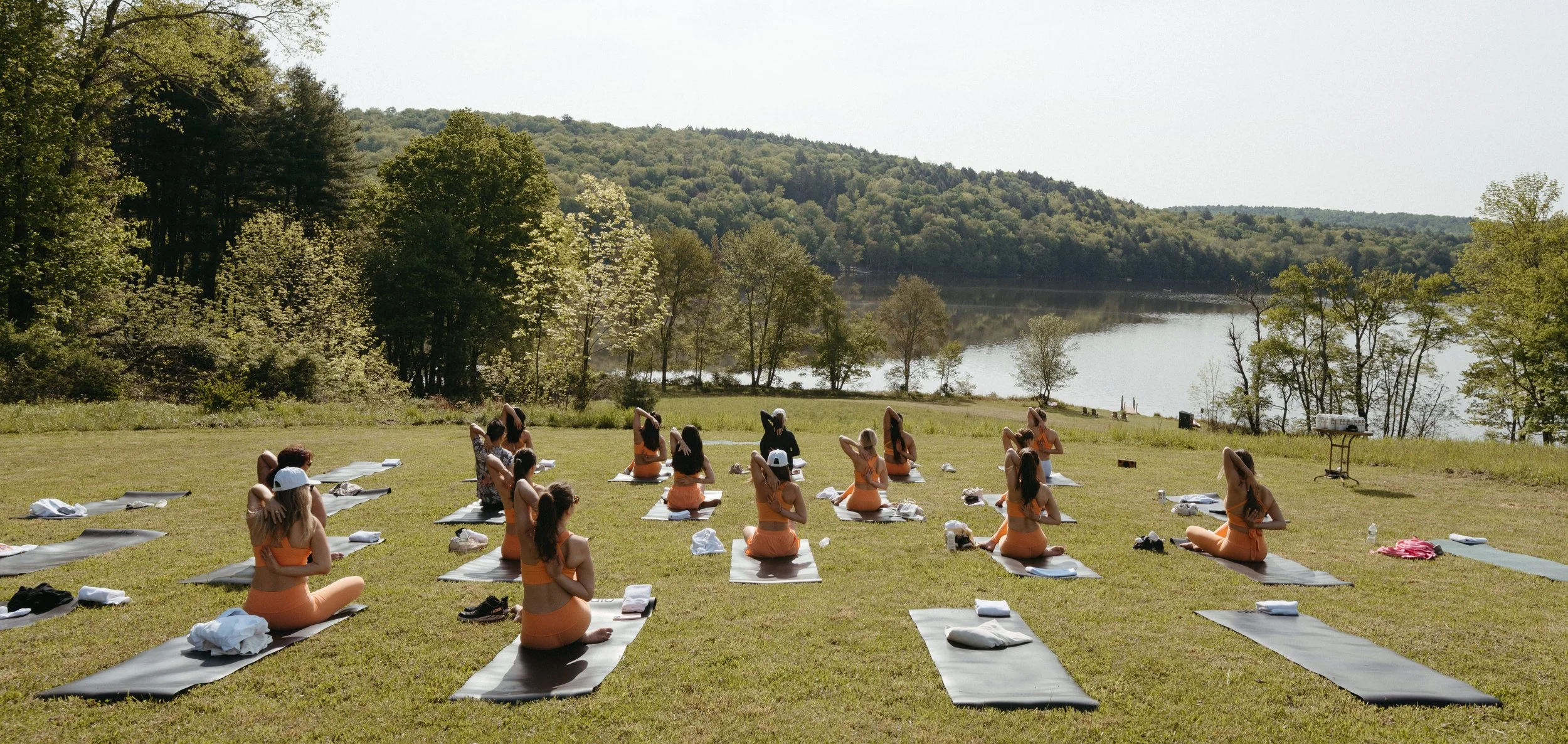 A group of people practicing yoga outdoors on a grassy field beside a lake, with trees and hills in the background on a sunny day.