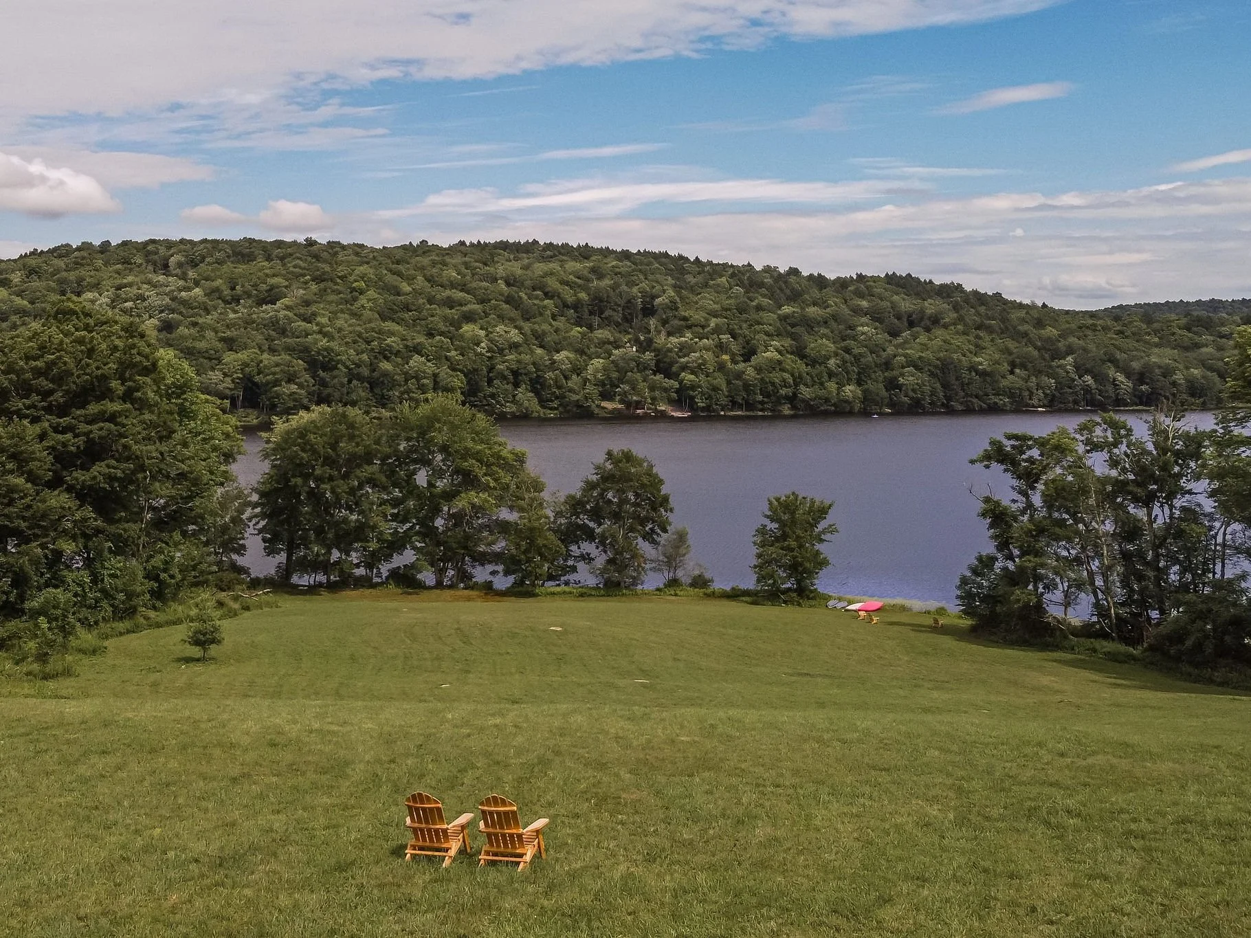 Two wooden chairs on green grass overlooking a lake with trees and hills in the background under a partly cloudy sky.