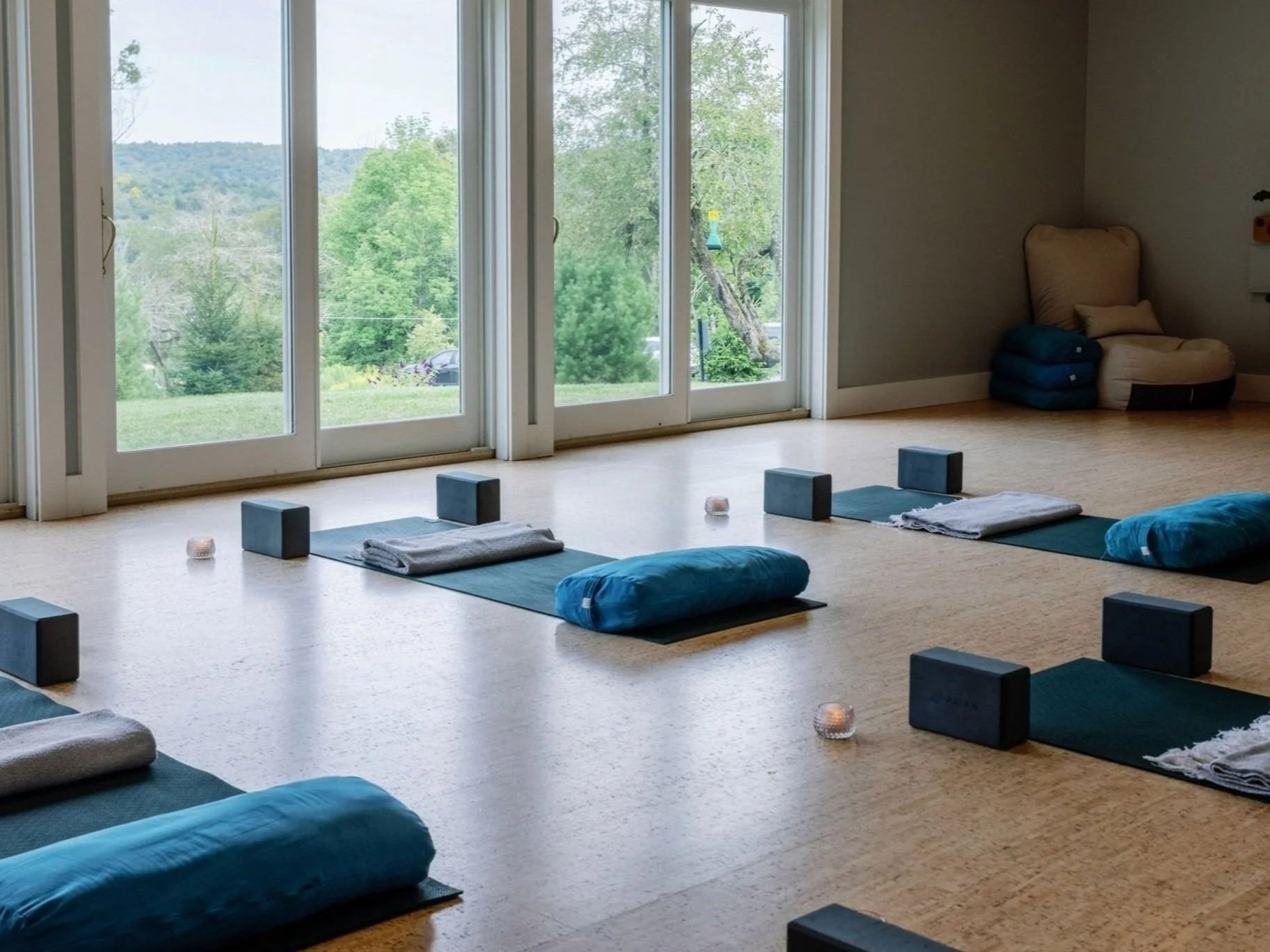 A serene yoga studio featuring large windows with a view of green trees and hills. The interior has mats, bolsters, and folded blankets arranged for a class. Candle lights add a calming ambiance.