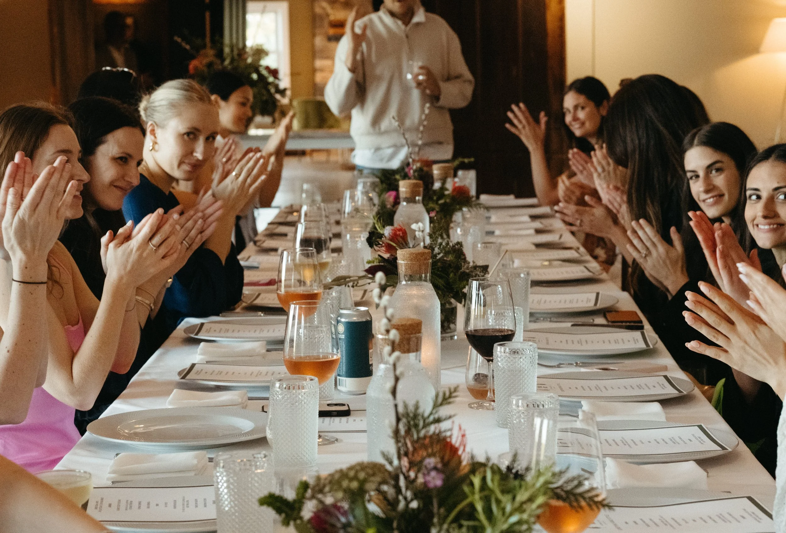 A group of women sitting at a long dining table, clapping, with a person standing at the head of the table holding a glass, in a warmly lit room with wooden walls and flowers on the table.