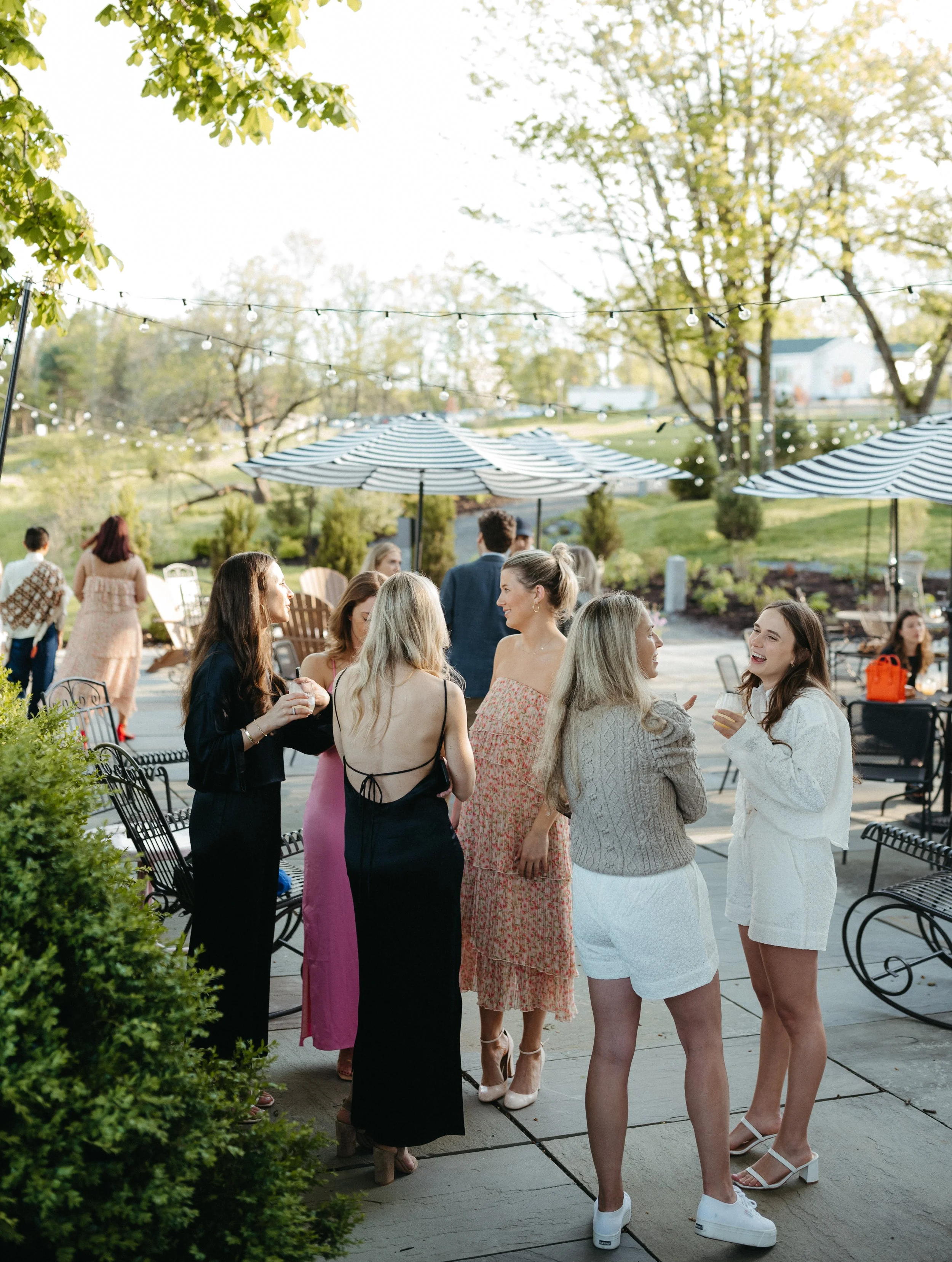 People gathered outdoors on a sunny day, socializing and enjoying drinks, with string lights and umbrellas in the background, and trees and houses beyond.