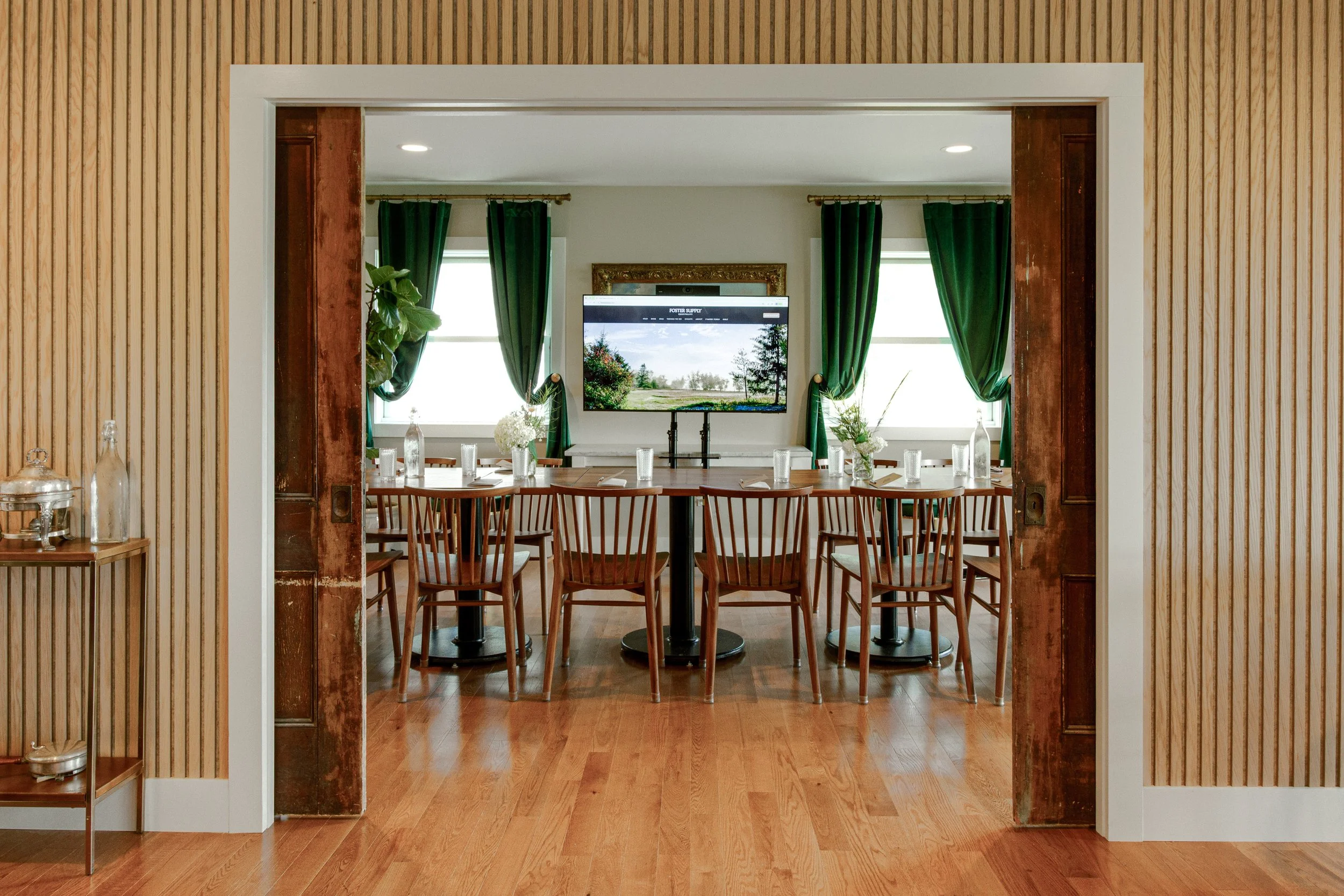 A dining room with a large wooden table and chairs, two large windows with green curtains, and a television mounted on the wall displaying a scenic view.