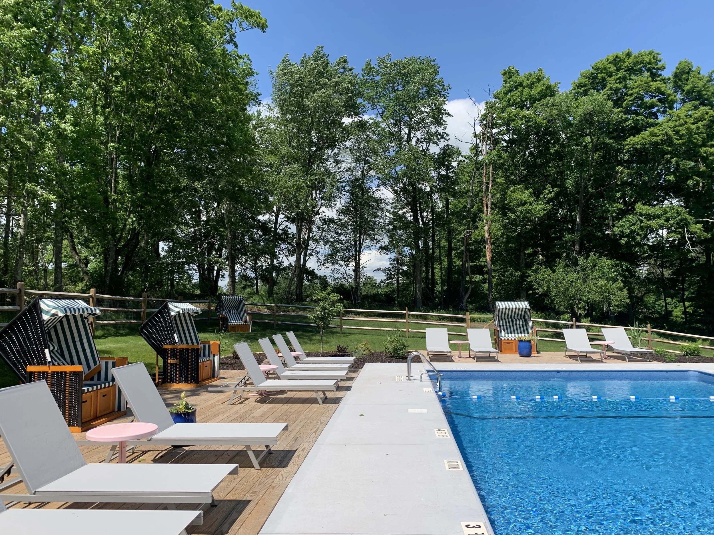 An outdoor swimming pool area with white lounge chairs, black and white striped chairs, pink and blue small tables, and a wooden deck. In the background, there are tall green trees and a bright blue sky with some clouds.