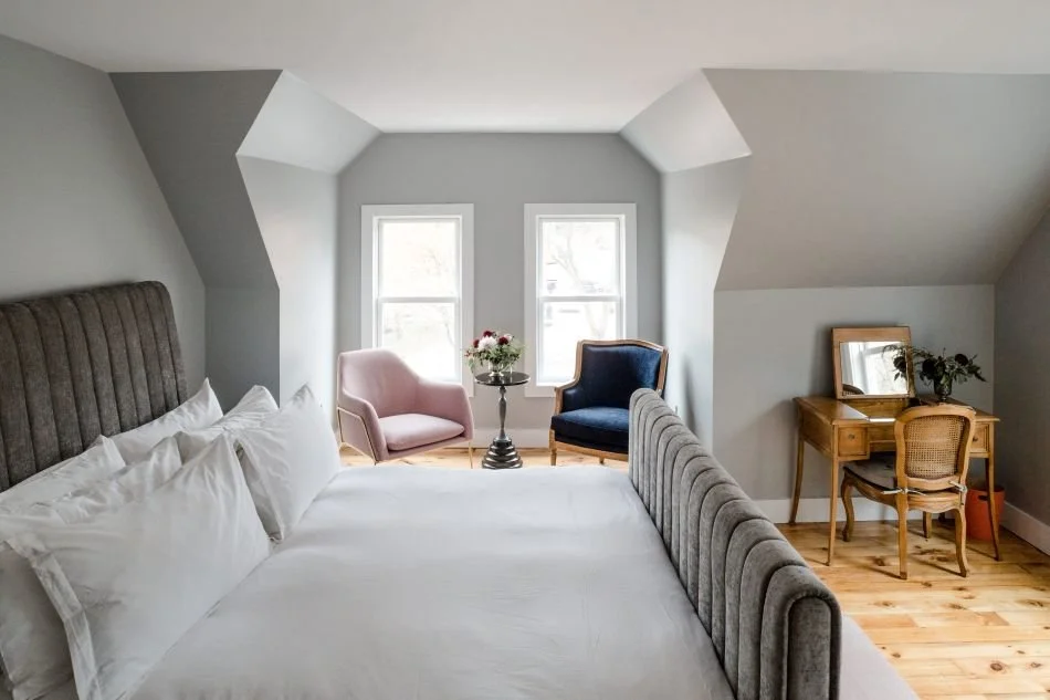 A cozy bedroom with a large bed, two pink and blue armchairs, a small table with flowers, and a vintage wooden dressing table with a mirror and a chair, in a room with two windows and hardwood floors.