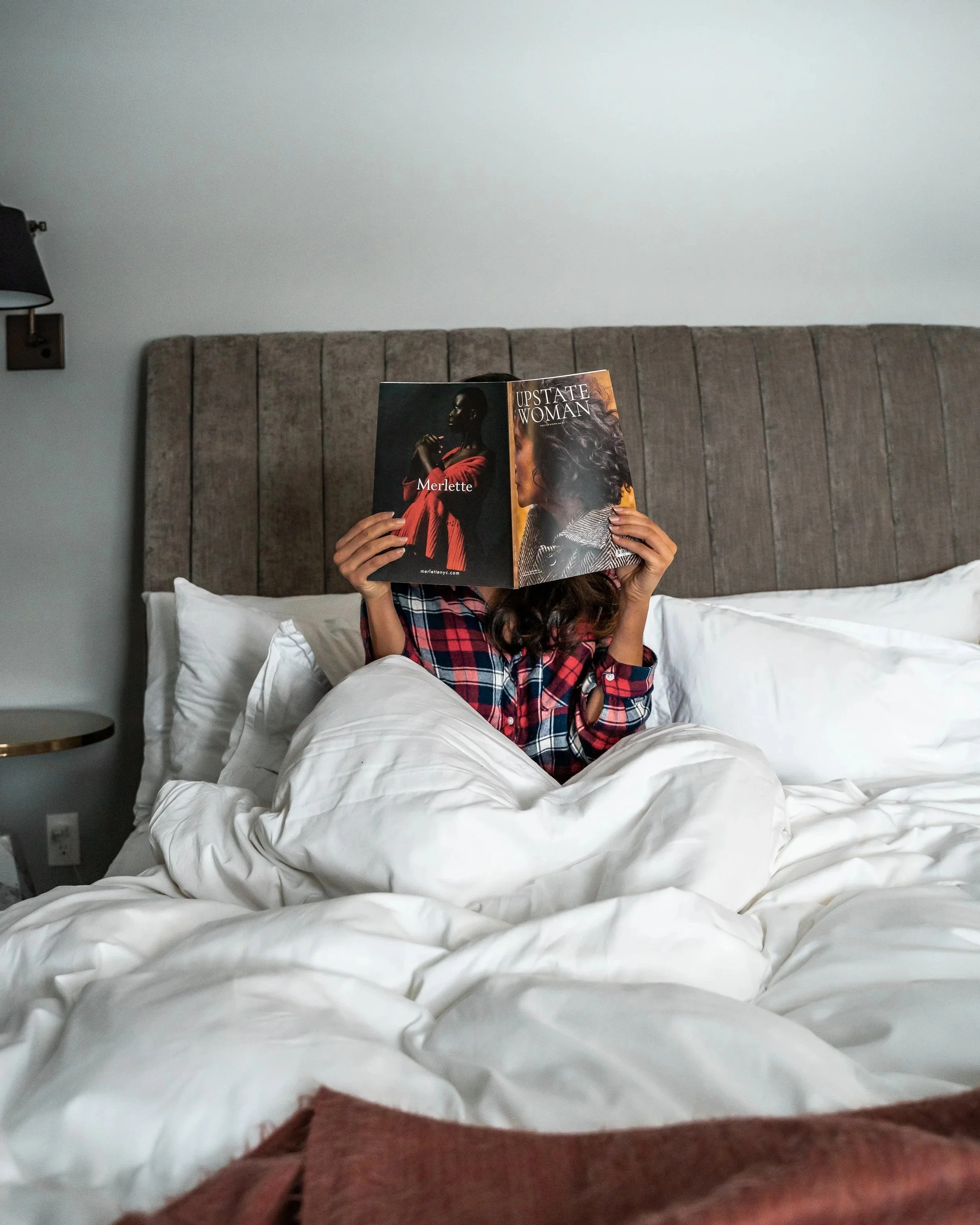 Person sitting in bed with white bedding, reading a magazine titled 'Upstate Woman' with another magazine inside, wearing a red and blue plaid shirt, against a gray headboard in a bedroom.