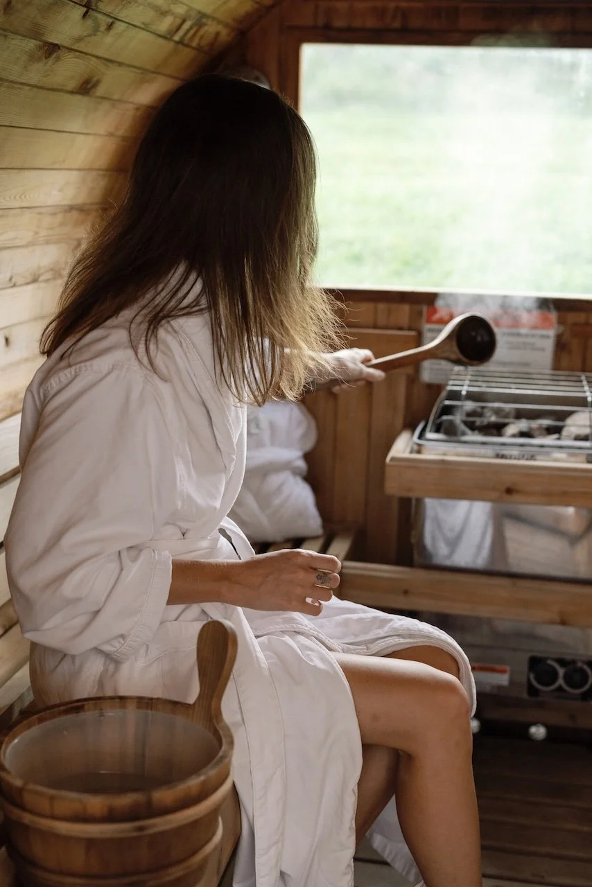 A woman sitting on a wooden bench in a sauna, stirring a bowl with a wooden spoon, with wood-paneled walls and a window showing greenery outside.