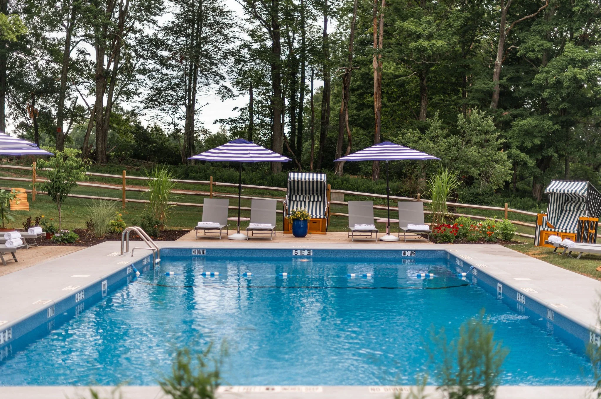 A swimming pool with blue water and depth markers, surrounded by a deck with lounge chairs and striped umbrellas, set against a backdrop of lush green trees.