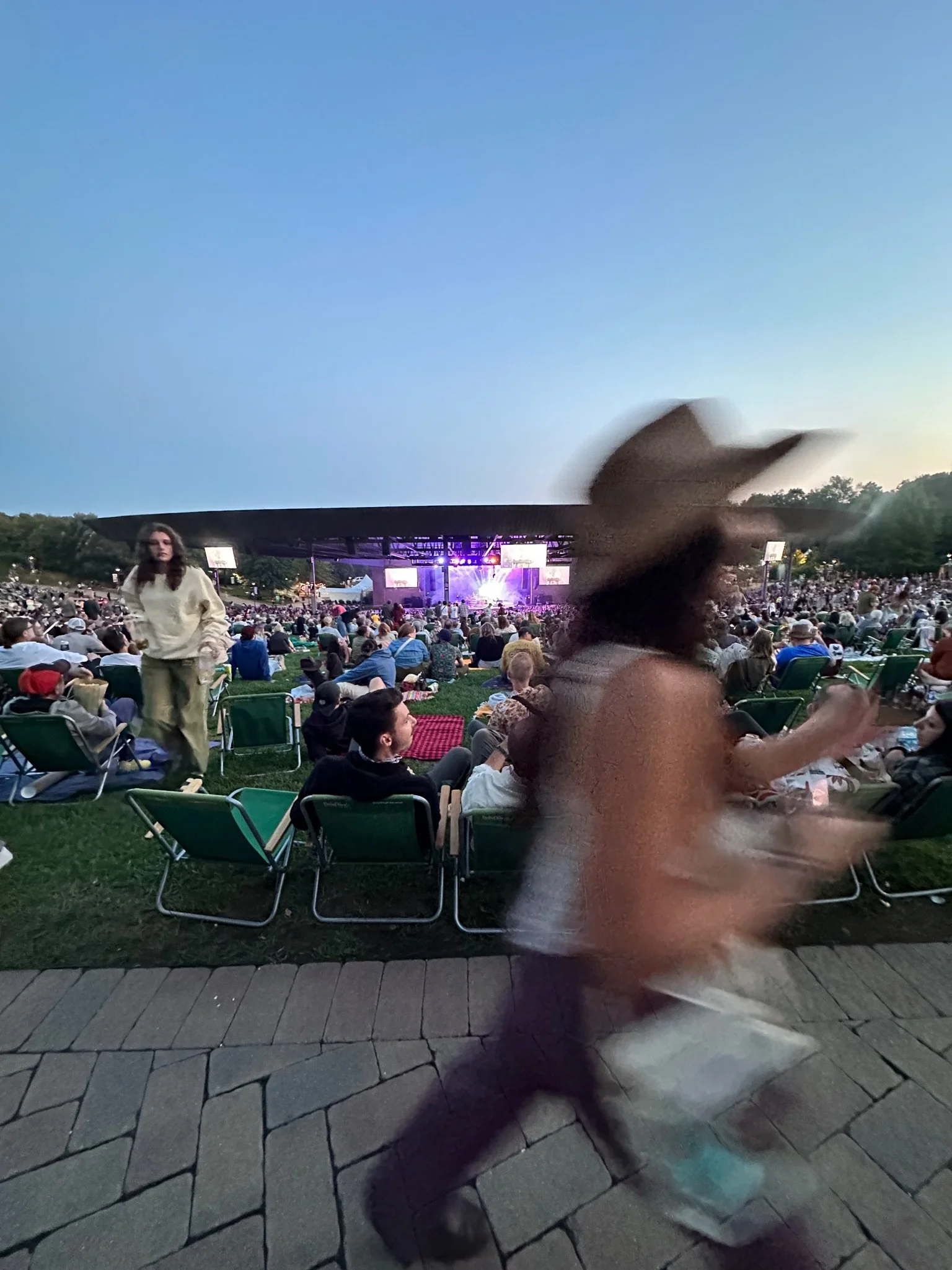 A large outdoor concert or festival at dusk with numerous people seated on grass and in chairs, a stage with bright lights, and a clear evening sky. In the foreground, a person wearing glasses and a white shirt and a woman in a beige jacket walk past, both slightly blurry.