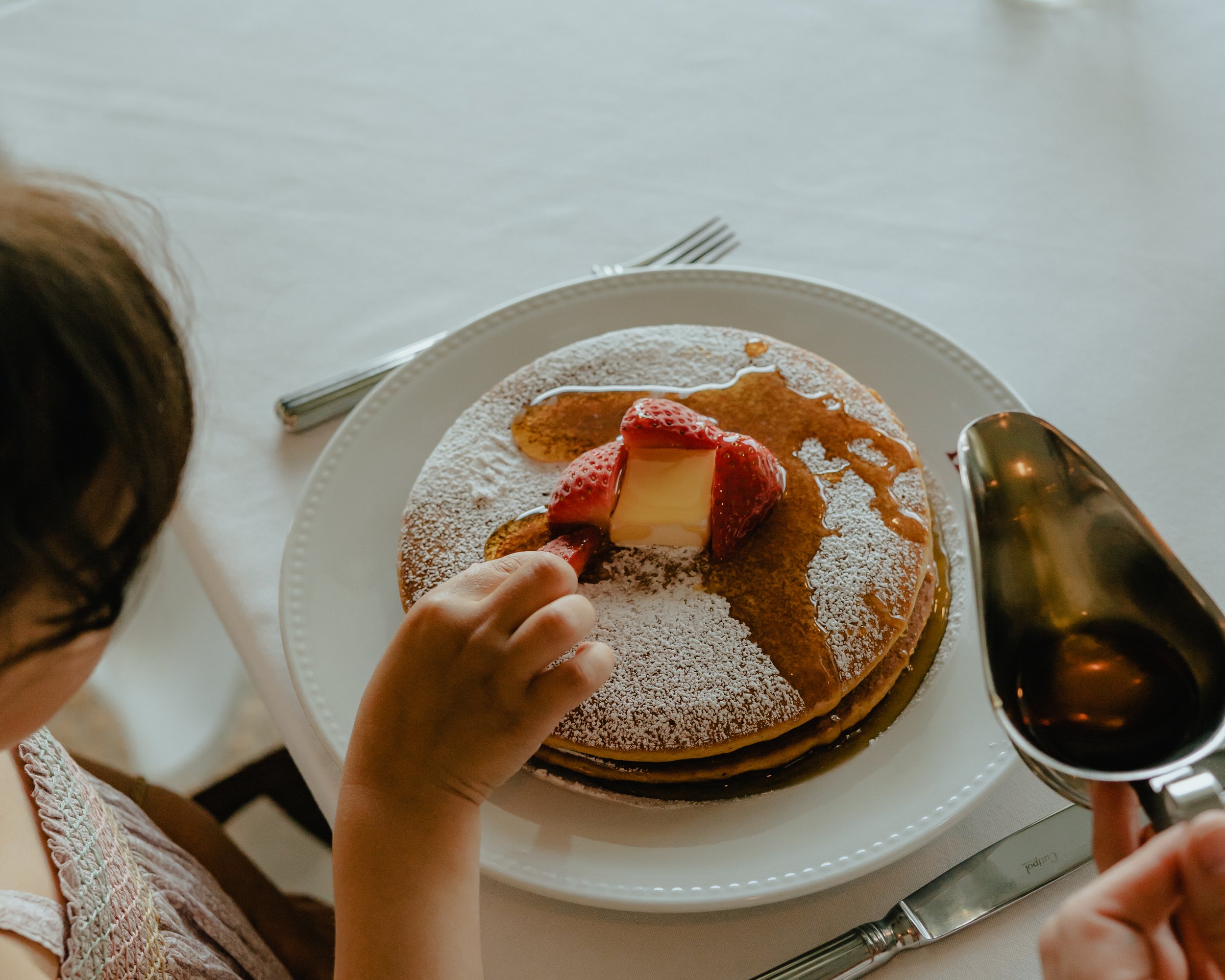 A child's hand reaching for a slice of strawberry-topped pancake on a white plate with powdered sugar, served with syrup in a gravy boat, on a table with a fork and knife.
