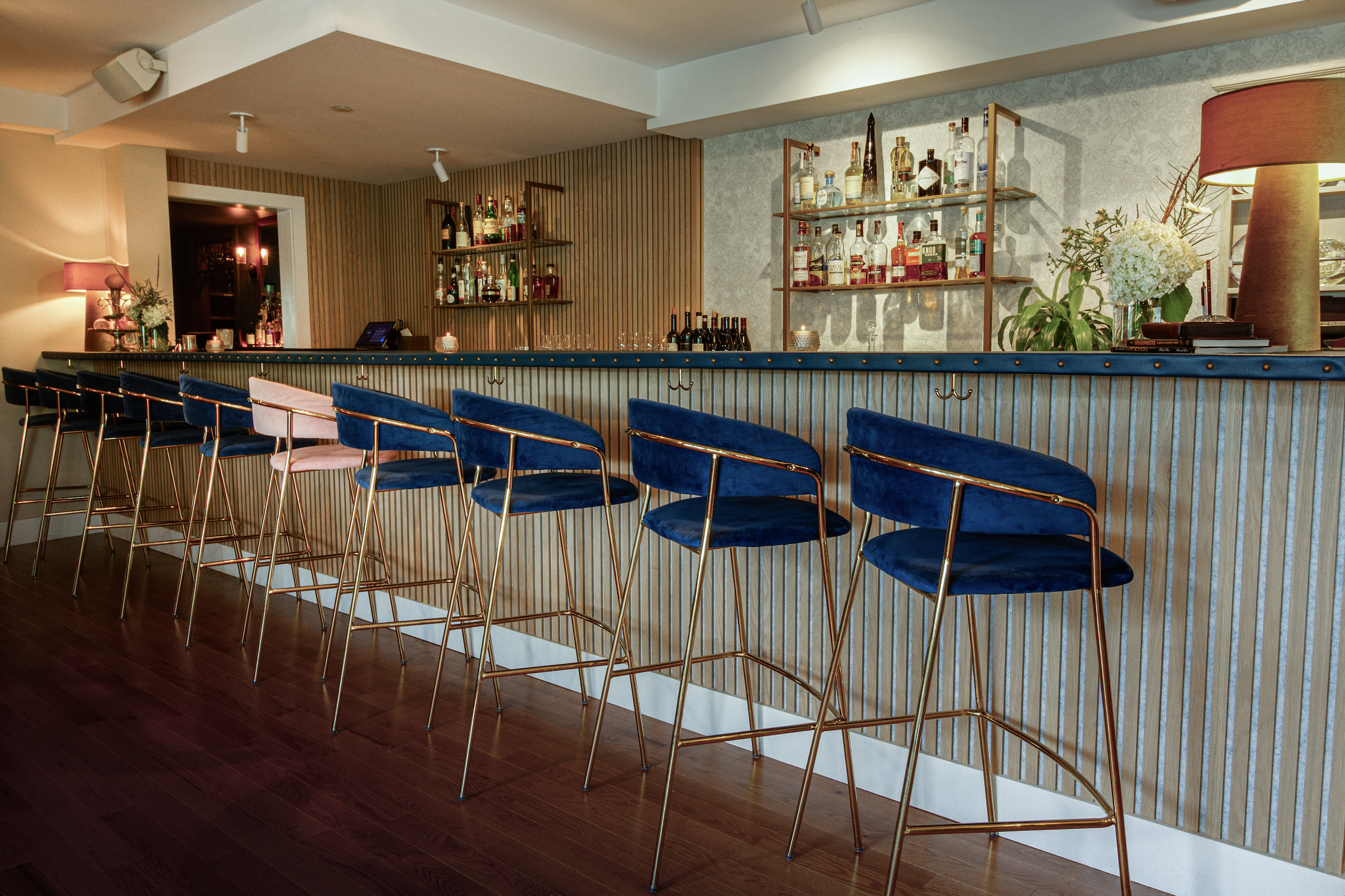 An interior view of a bar with a row of blue velvet bar stools with gold legs, a countertop with bottles of alcohol, shelves with more bottles, and decorative lamps and flowers.