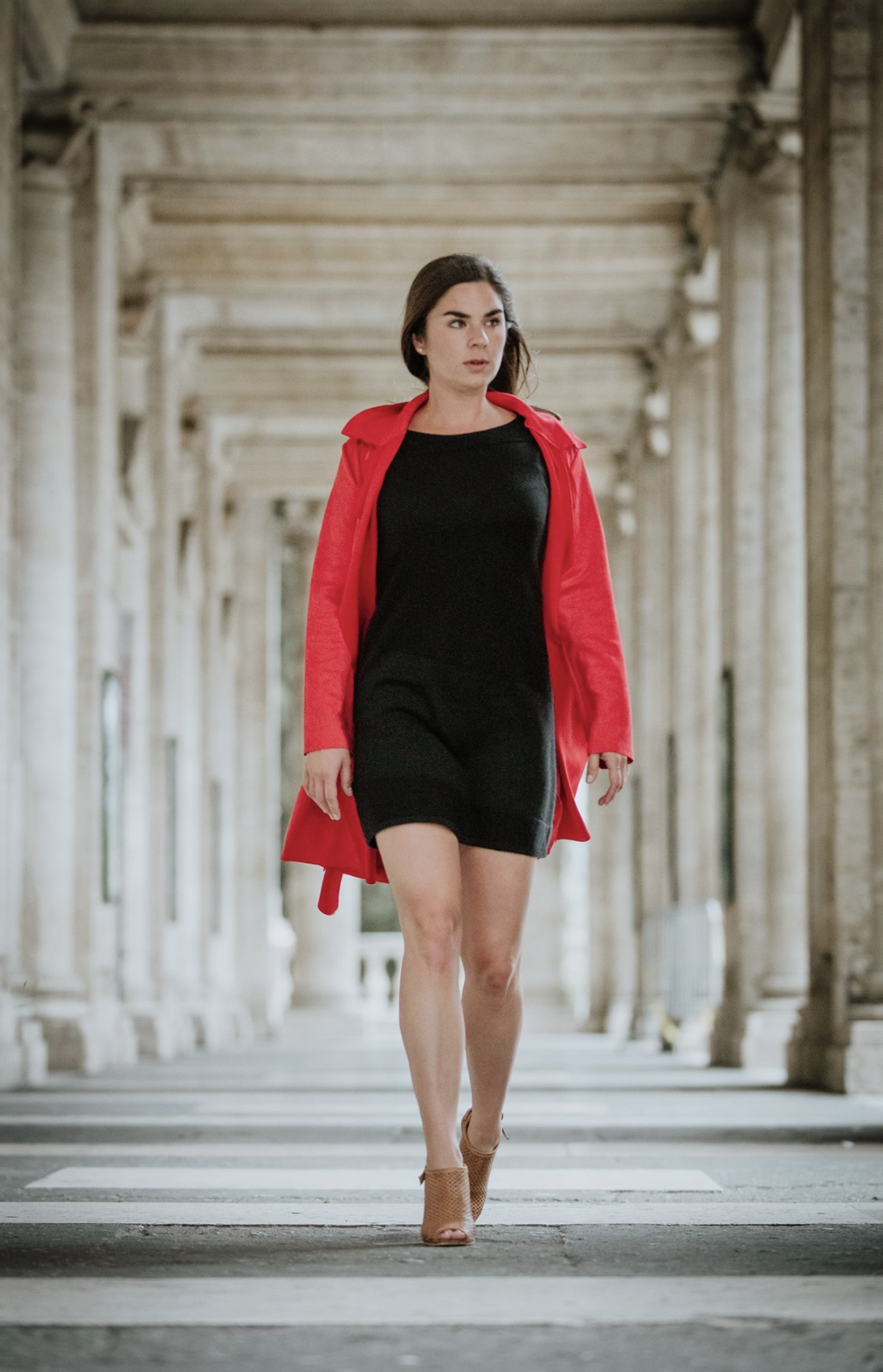 A woman walking in an outdoor corridor with stone columns, wearing a black dress, tan high heels, and a red coat.