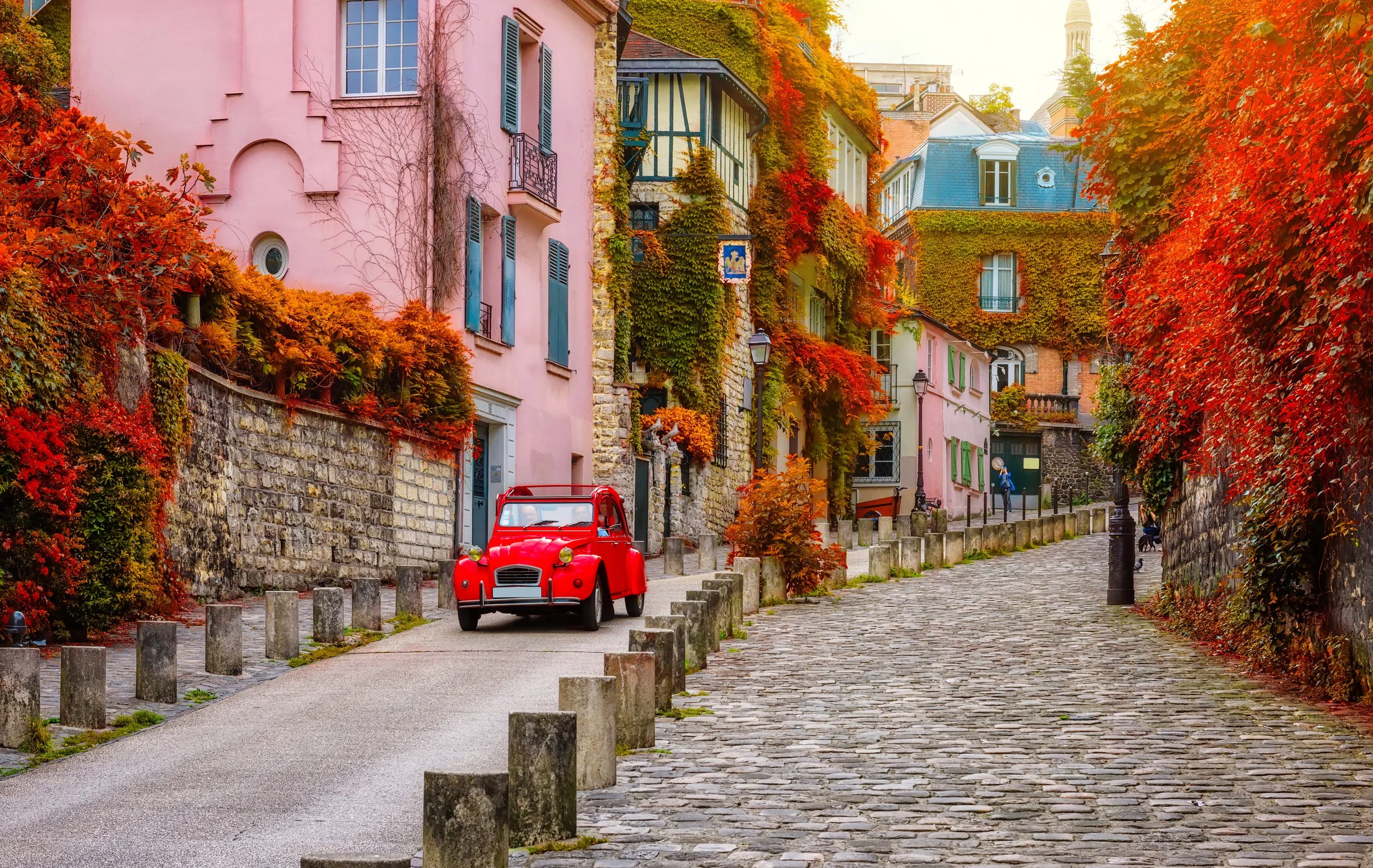 A cobblestone street lined with colorful houses covered in autumn foliage, with a red vintage car parked on the side and a person walking with a dog.
