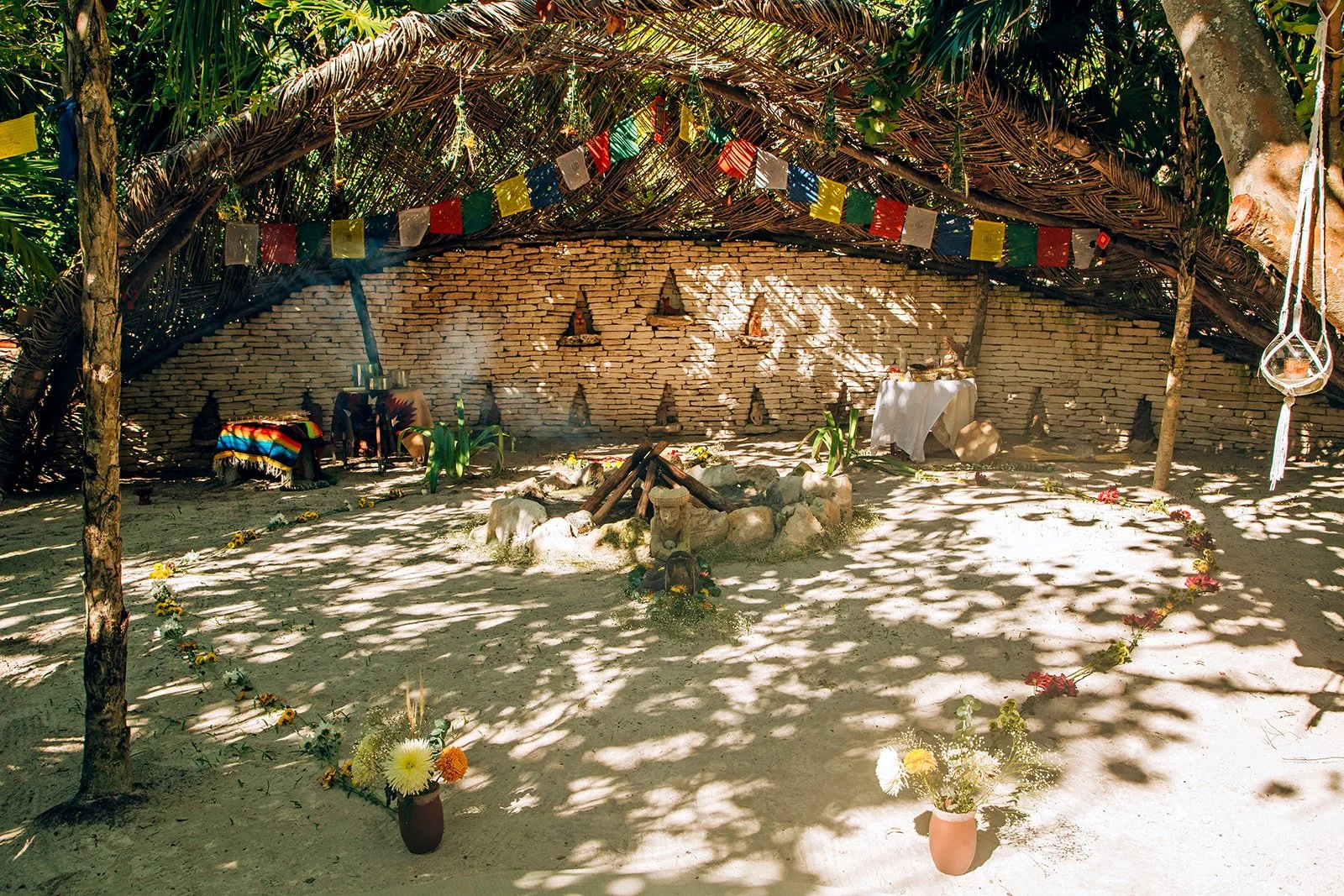 A shaded outdoor space with a thatched roof, decorated with colorful paper flags, set up for a small celebration or ceremony. There are flower arrangements, a small altar with statues, and a fire pit in the center surrounded by rocks.