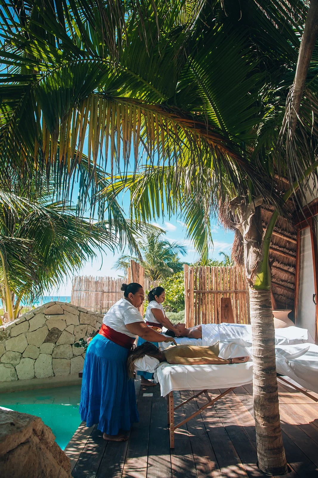 Two women getting massages outdoors near palm trees, with a thatched roof structure and bamboo fence, in a tropical setting.
