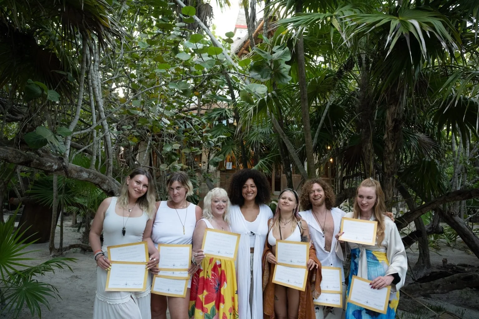 Group of seven women standing outdoors in a tropical setting, holding certificates and smiling.