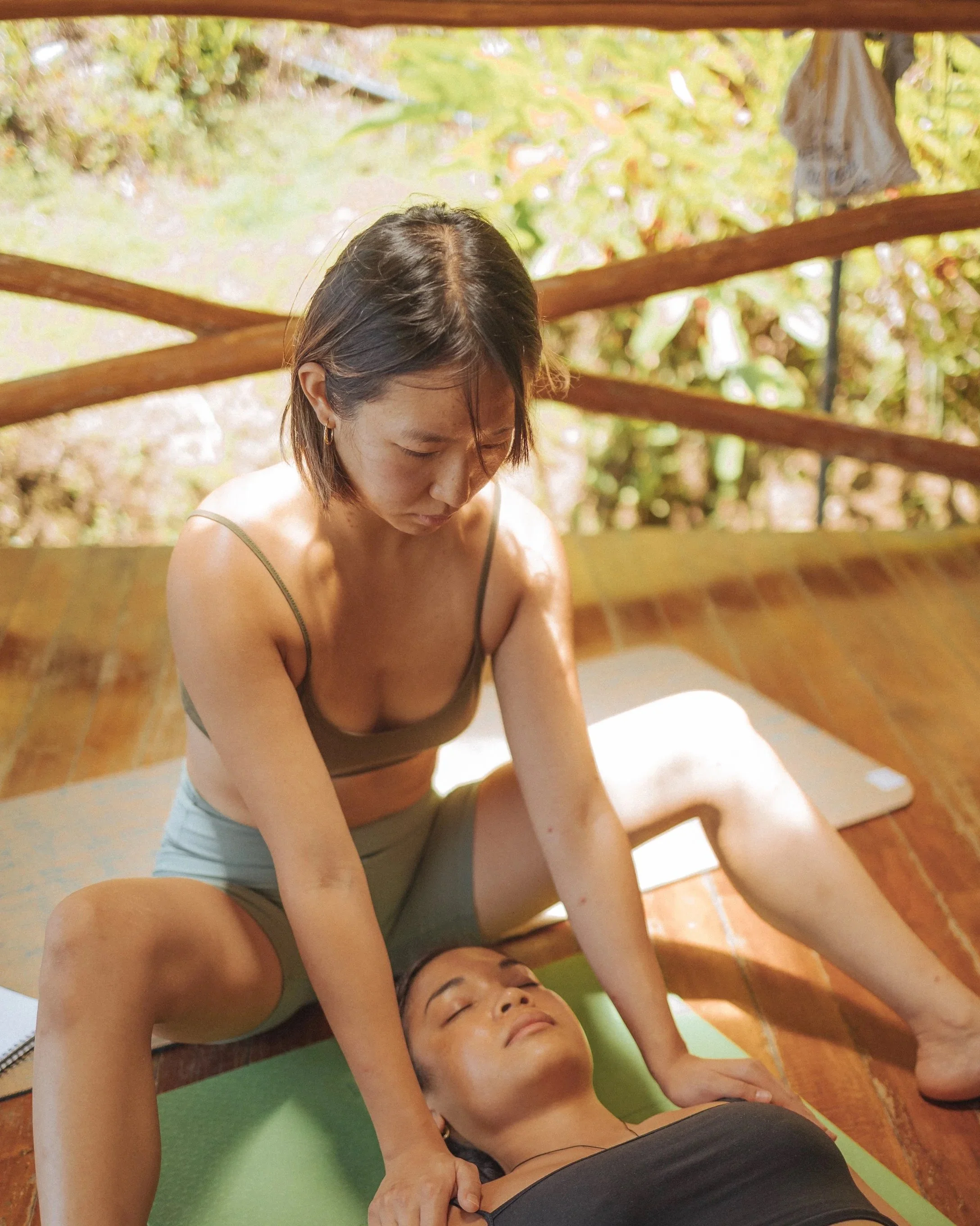 A woman giving a shoulder massage to another woman lying on a yoga mat on a wooden floor, with a background of trees and outdoor scenery.