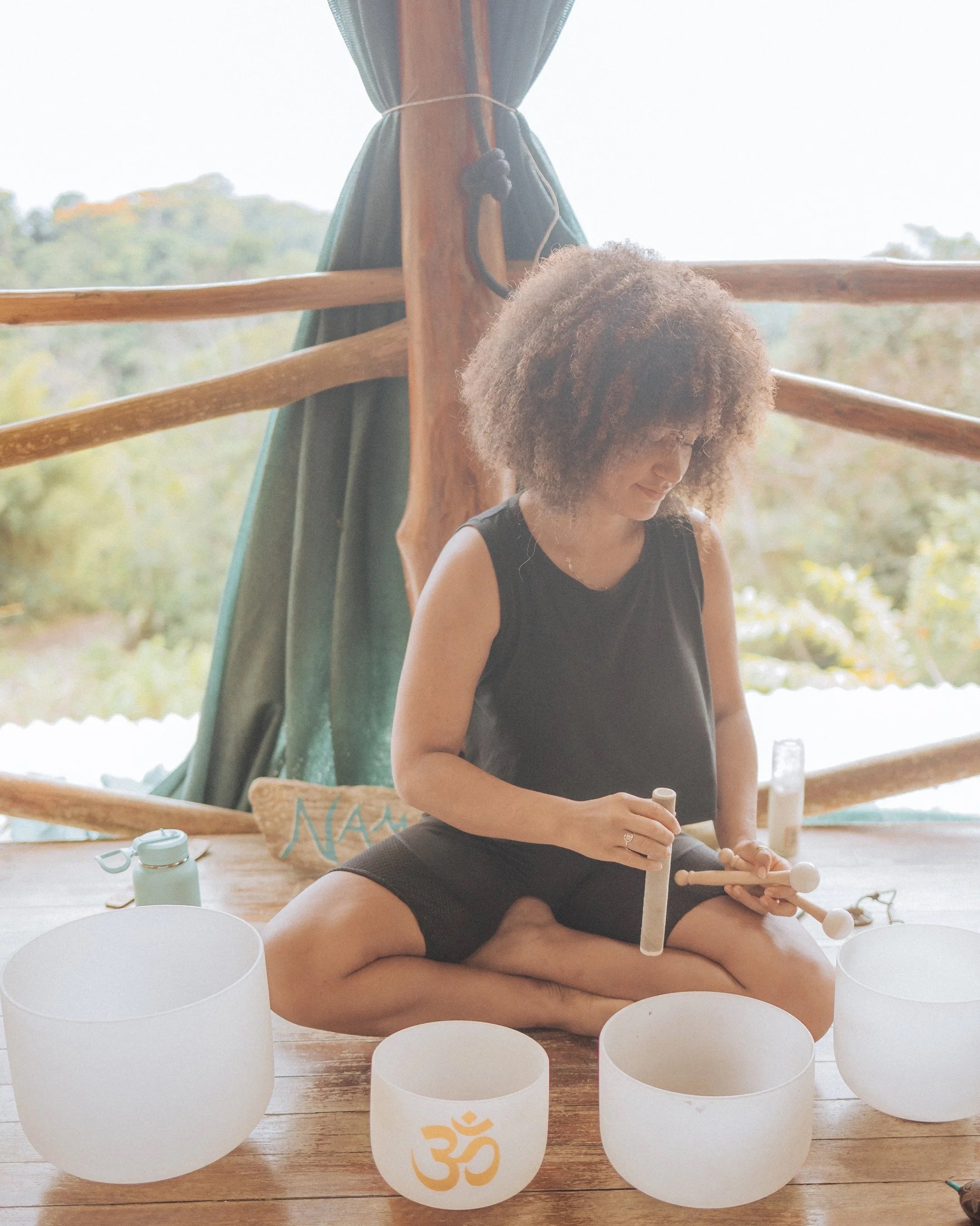 A woman with curly hair sitting cross-legged on a wooden floor, playing crystal singing bowls. She is inside a rustic wooden structure with a window showing a natural landscape outside.