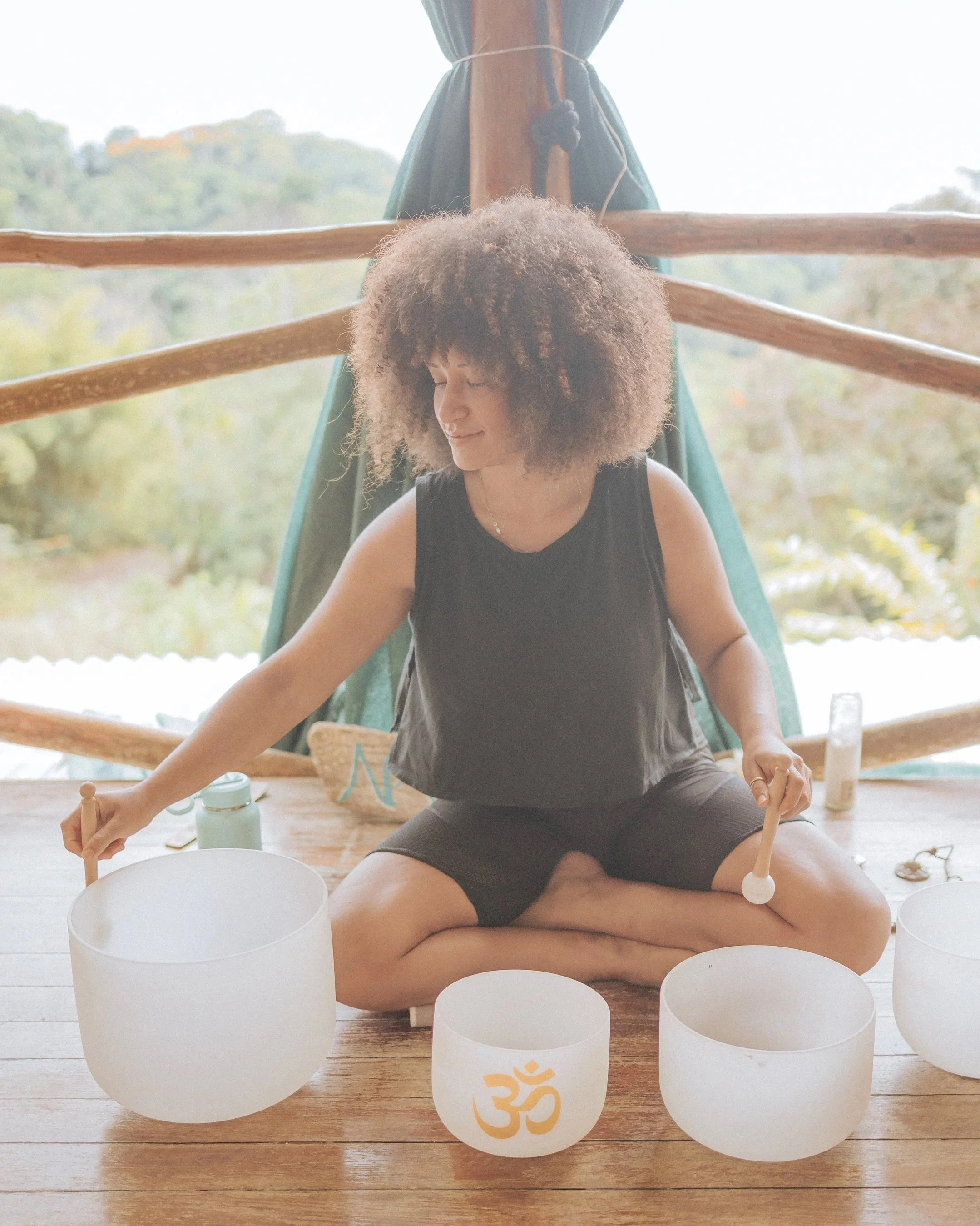 A woman sitting cross-legged on a wooden floor playing crystal singing bowls, with a window showing a green outdoor landscape behind her.