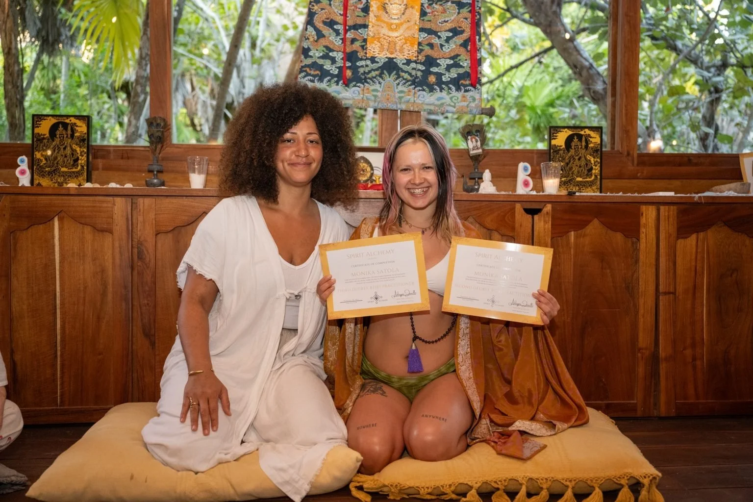Two women sitting on a yellow cushion, smiling, holding certificates at a spiritual or meditation event with a wooden altar and decorative items behind them.