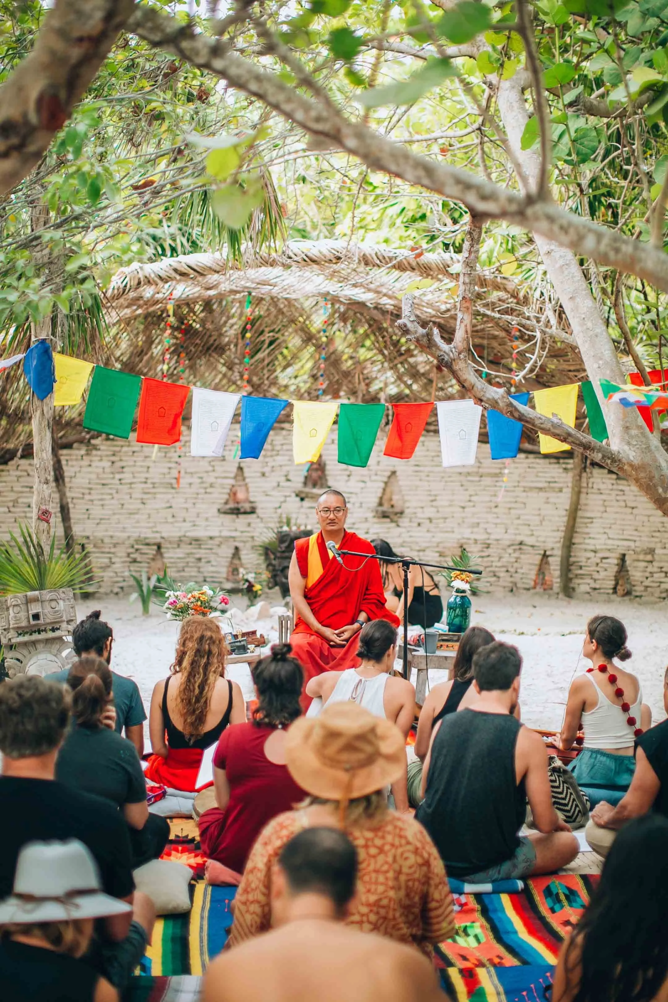 A man in red robes sitting cross-legged and speaking into a microphone to a seated audience under a tent-like structure with colorful flags, surrounded by greenery and trees.