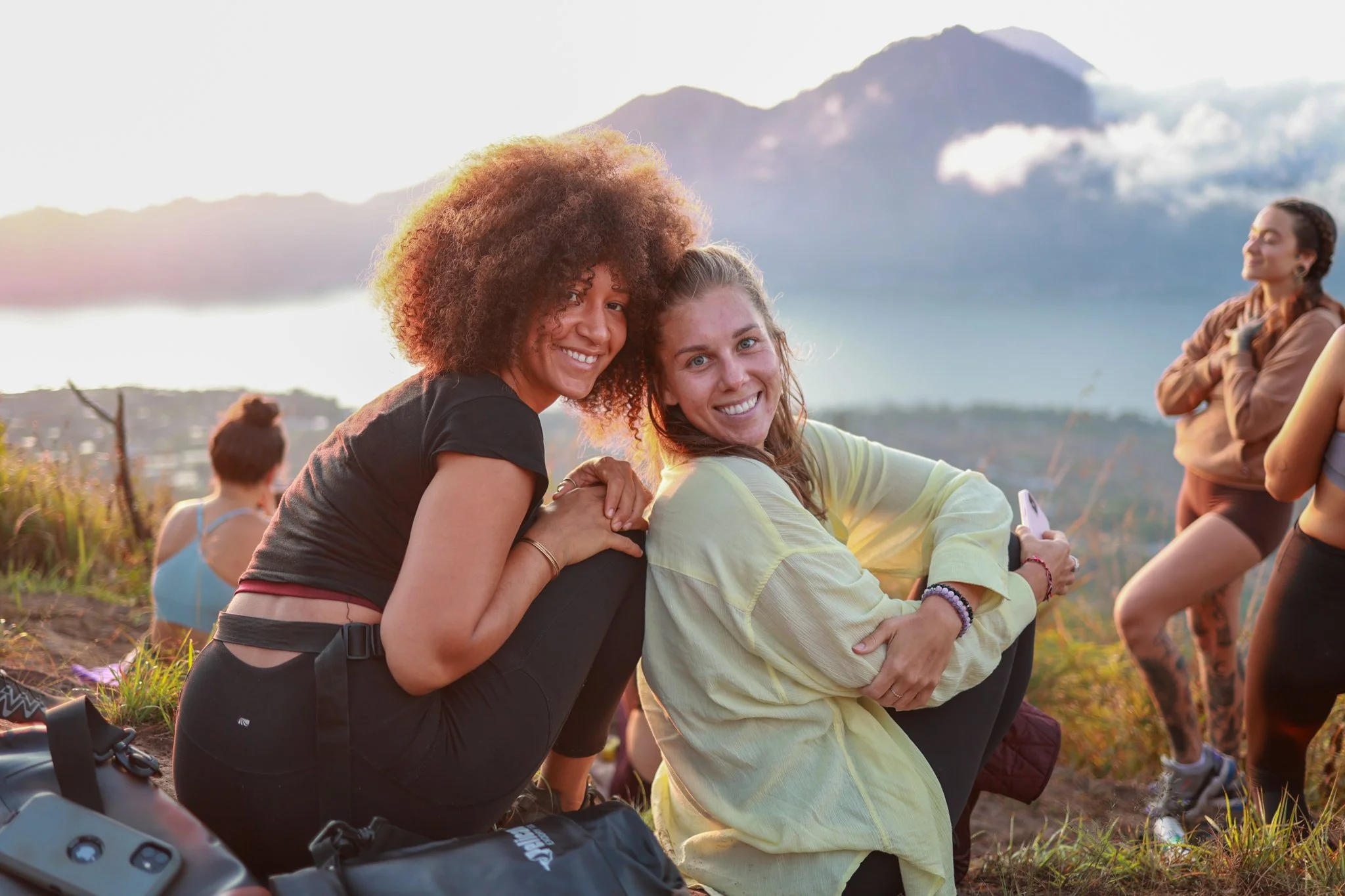 Two women sitting closely together on a grassy hill, smiling at the camera. In the background, a mountain and a cloudy sky are visible, with other women standing and sitting nearby.