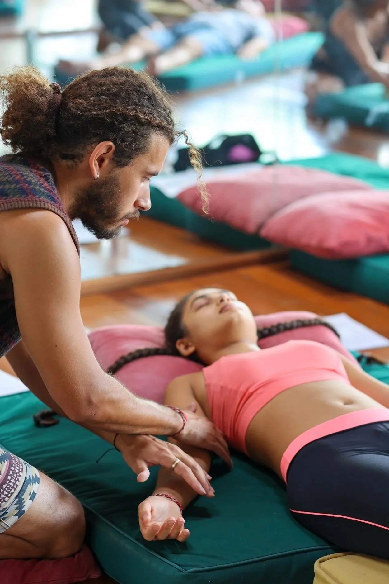 A person performs CPR on a young woman lying on a mattress with pink pillows in a room with multiple mattresses and pillows.