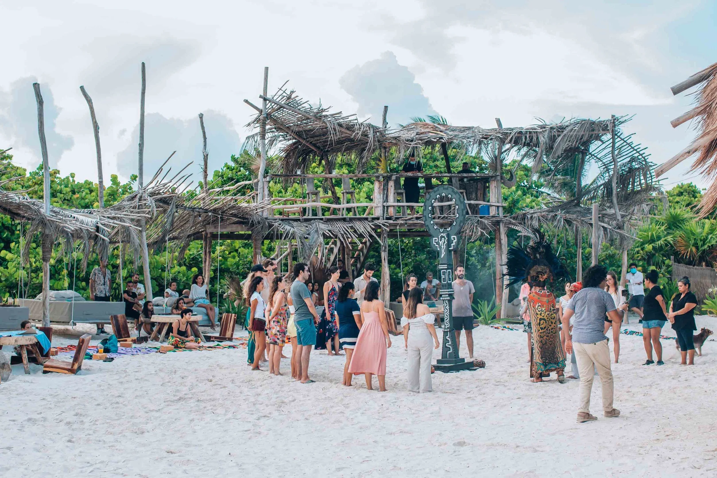 Group of people participating in a cultural or spiritual ceremony on a sandy beach with a rustic wooden hut in the background, surrounded by green tropical foliage.