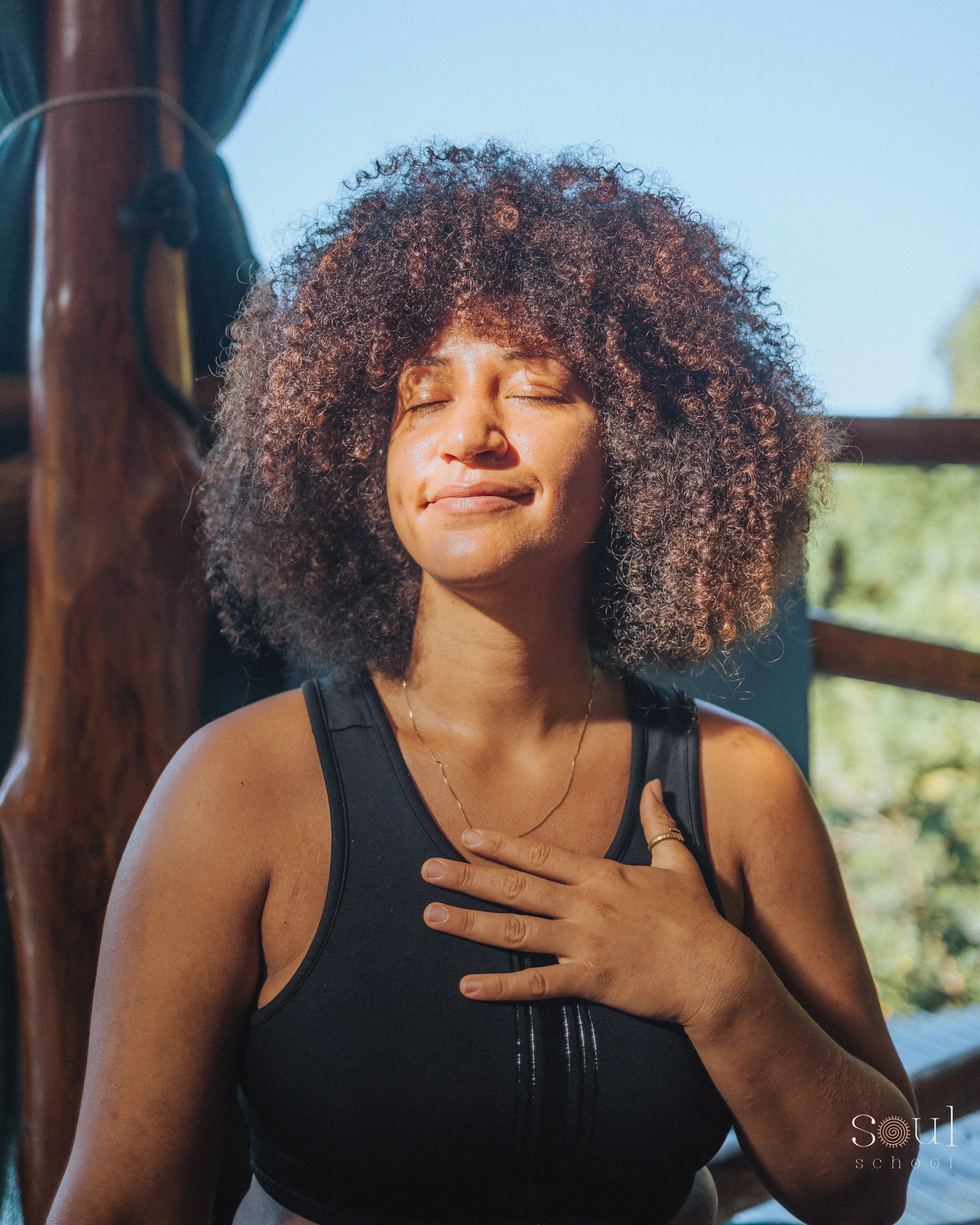 A woman with curly hair and a black tank top, eyes closed, smiling peacefully, with her hand on her chest, in a sunlit room with wooden beams and a window showing greenery outside.