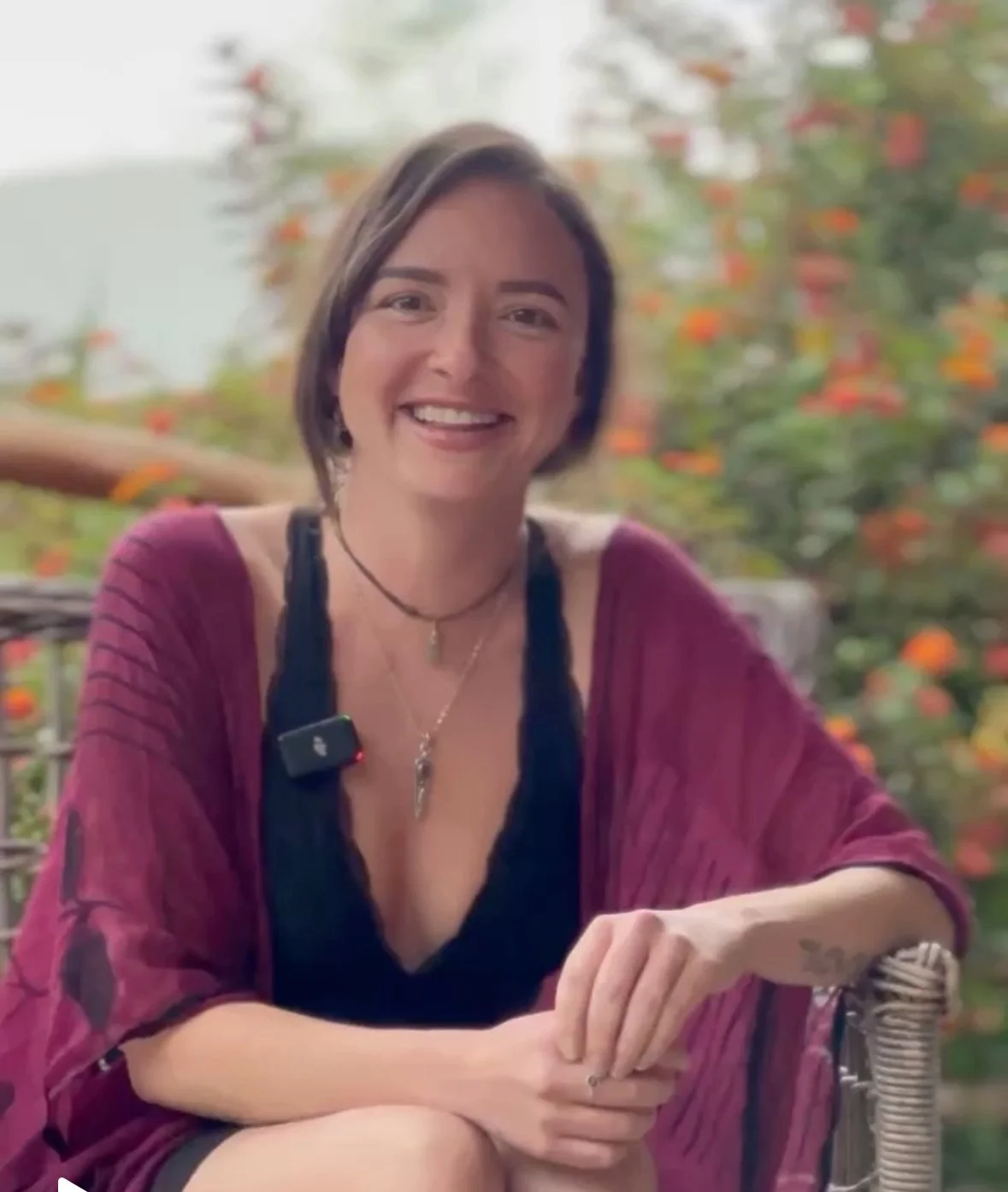 A woman with short brown hair smiling at the camera, sitting outdoors on a chair, with a blurred background of orange flowers and greenery.