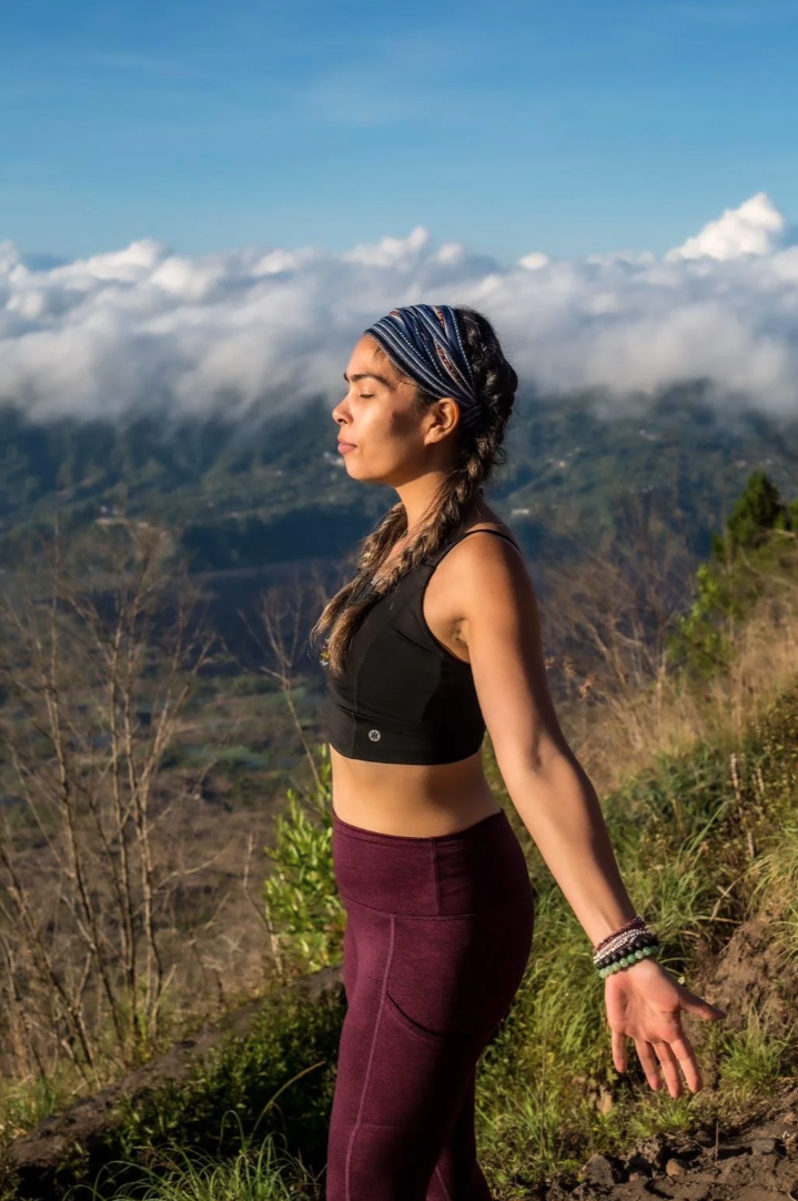 Costa Rica Yoga & Reiki Immersion program. A woman practicing yoga outdoors on a mountain trail, with her eyes closed and arms relaxed at her sides, surrounded by scenic hills and clouds.