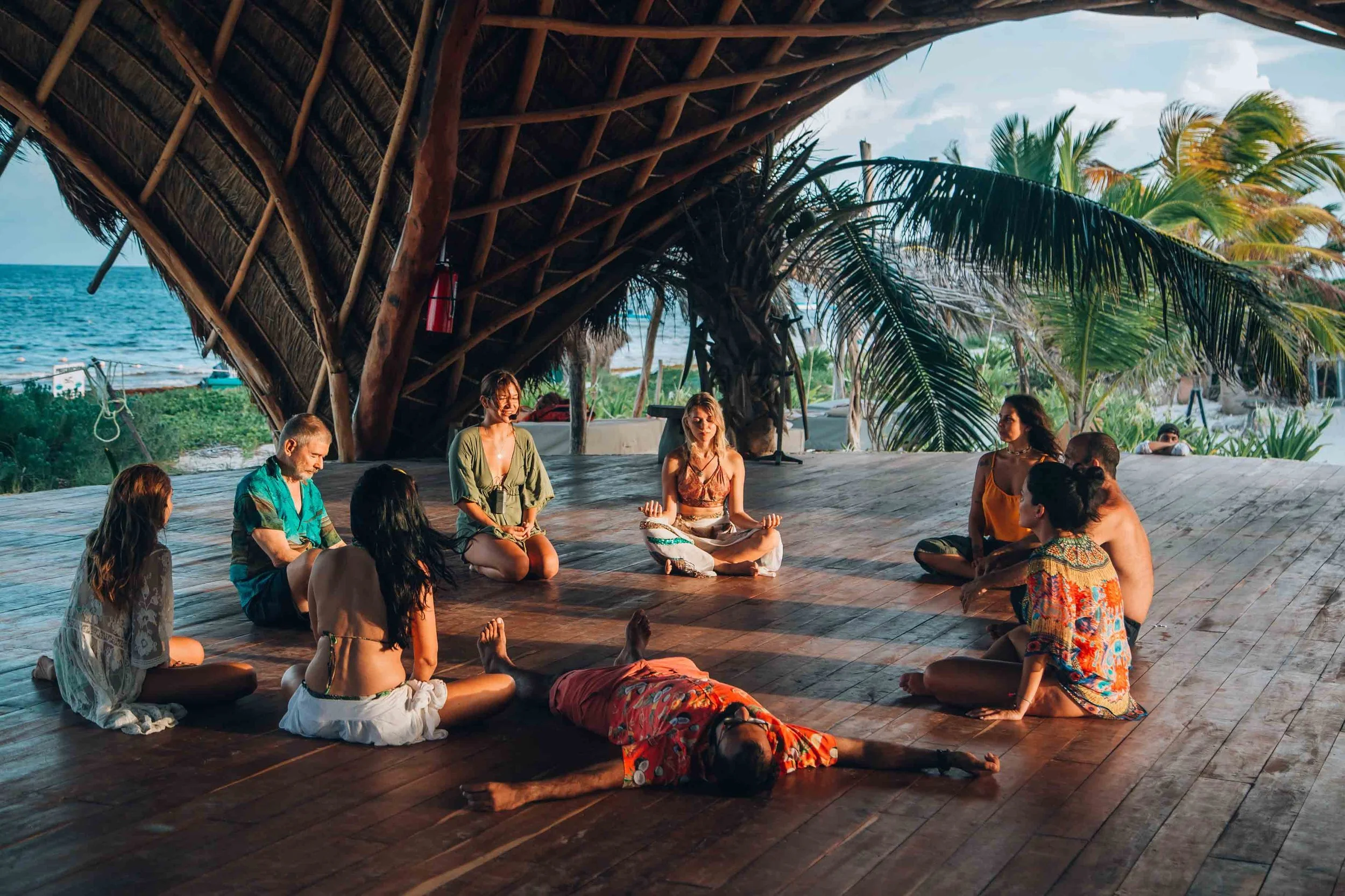 People practicing yoga in a circle on a wooden platform under a thatched roof with palm trees and the ocean in the background