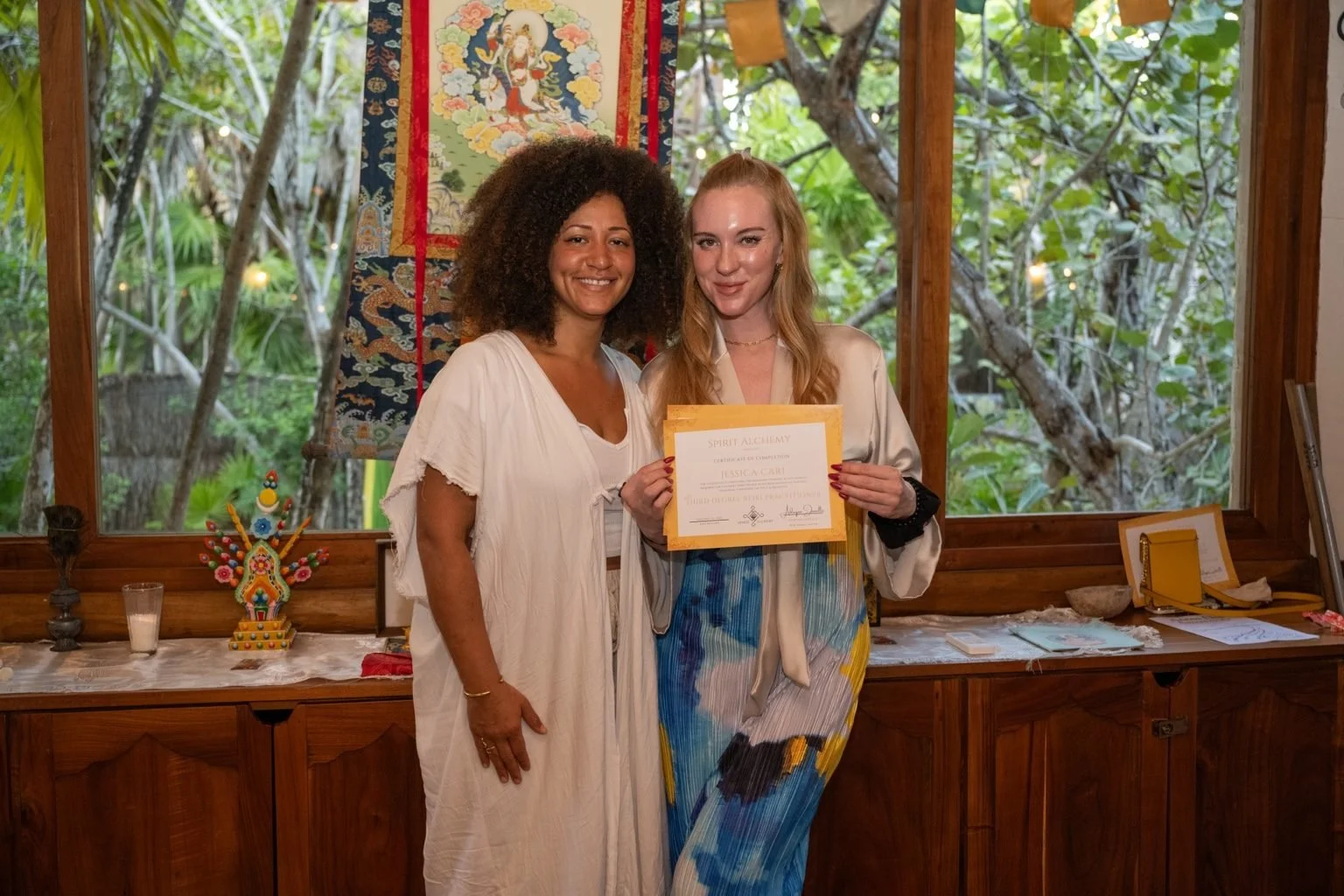 Two women standing inside, one holding a certificate, with a decorative tapestry and a window showing greenery behind them.