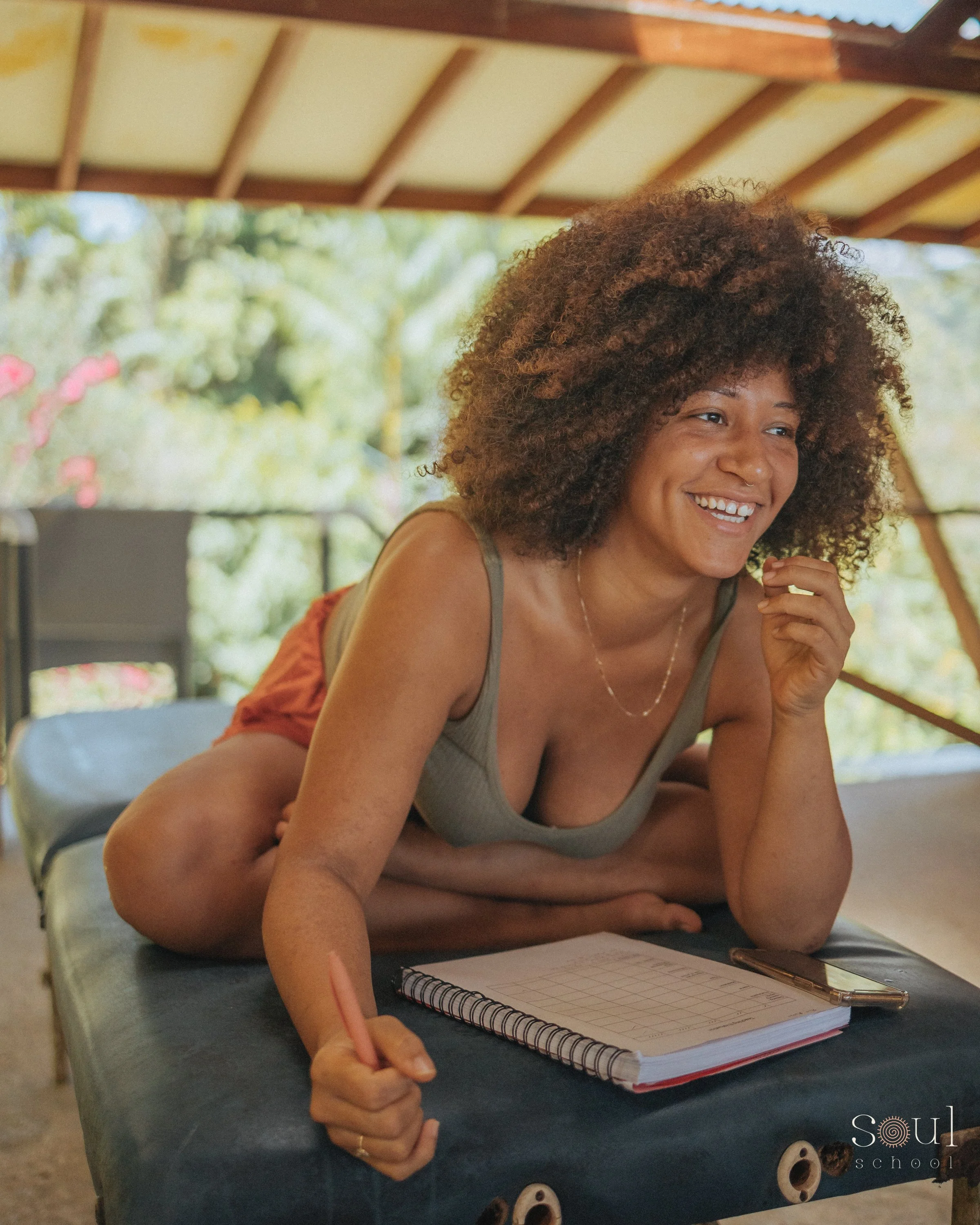 Woman with curly hair lying on her stomach on a massage table, writing in a notepad, with a smile and relaxed expression, in a natural outdoor setting.