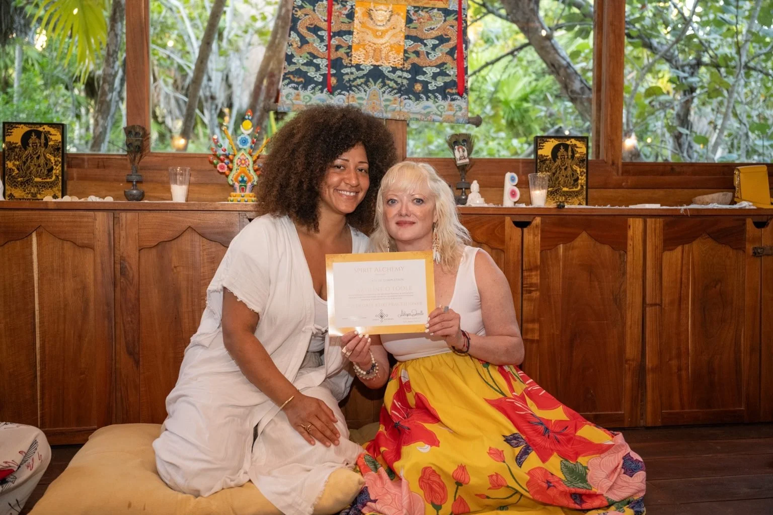 Two women sitting on a cushioned seat, holding a certificate. One woman has dark curly hair and dark skin, wearing a white outfit. The other woman has blonde curly hair and light skin, wearing a white top and a colorful floral skirt. They are inside 