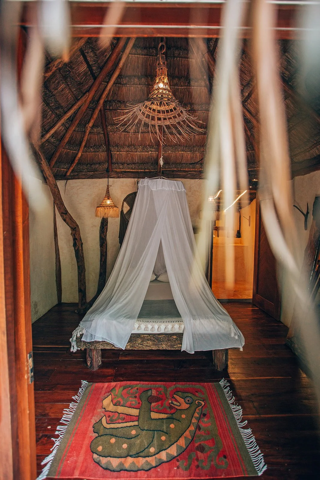 Interior of a rustic bedroom with a canopy bed draped in white fabric, wooden furniture, hanging woven lampshades, and a colorful rug with a lizard design on a wooden floor.