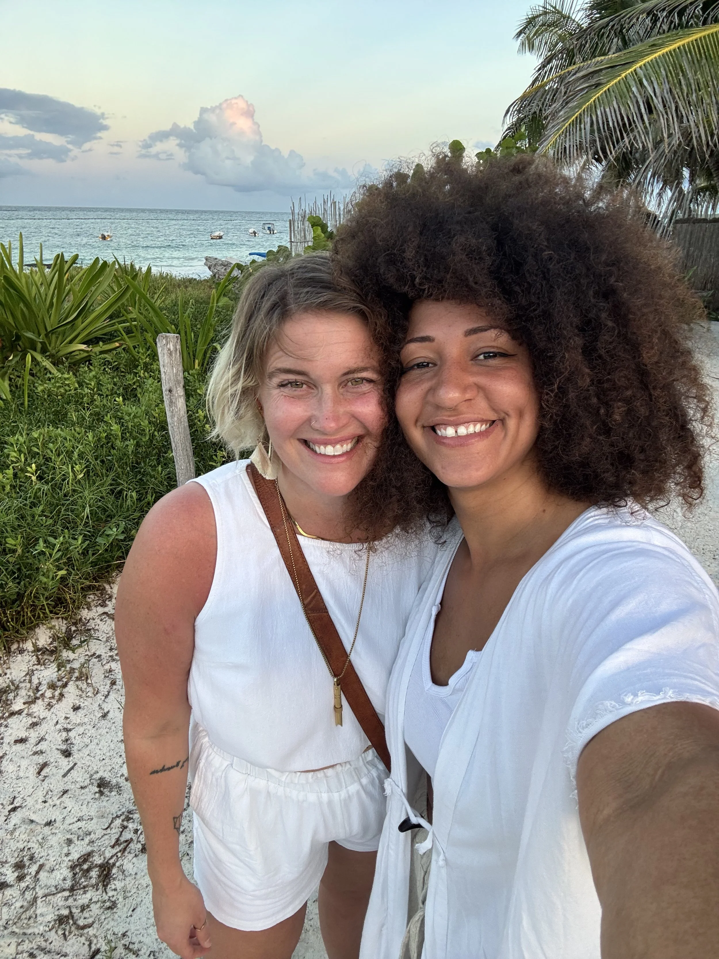 TTulum Reiki Certification Retreat experience. wo women smiling on a beach, with ocean, boats, and palm trees in the background during sunset.