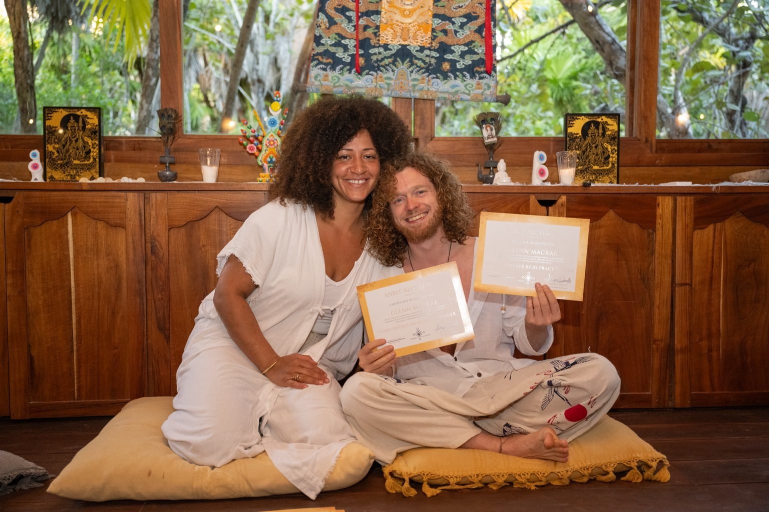 A woman and a man sitting on cushions on the floor, holding certificates, smiling. The woman has dark curly hair and wears a white outfit, the man has red curly hair and beard, dressed in white with patterned pants, inside a wooden room with large wi