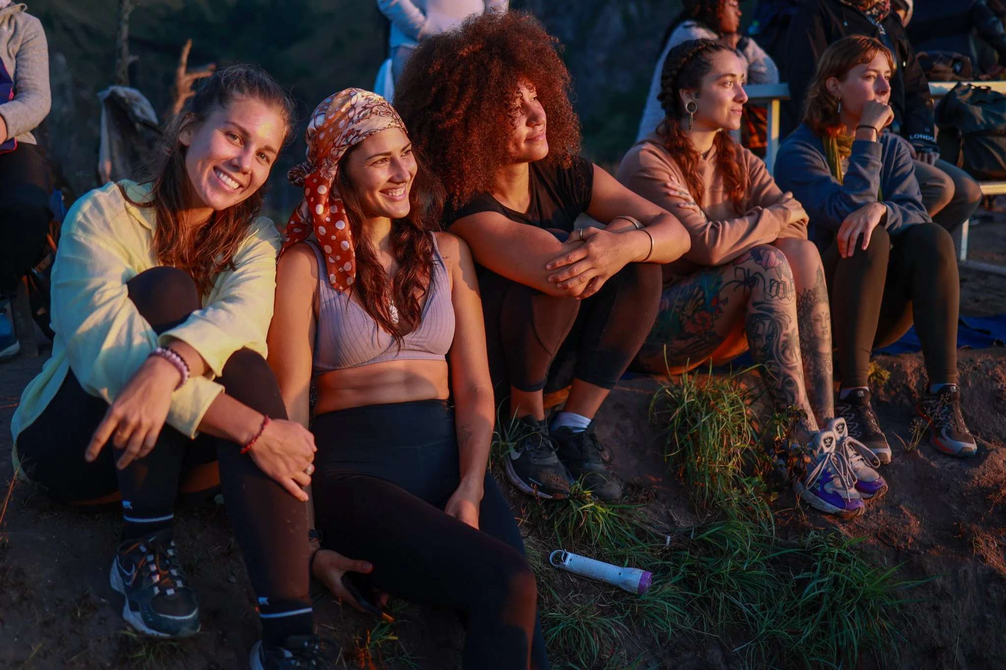 Group of young women sitting on the ground outdoors during sunset, watching a performance or event.