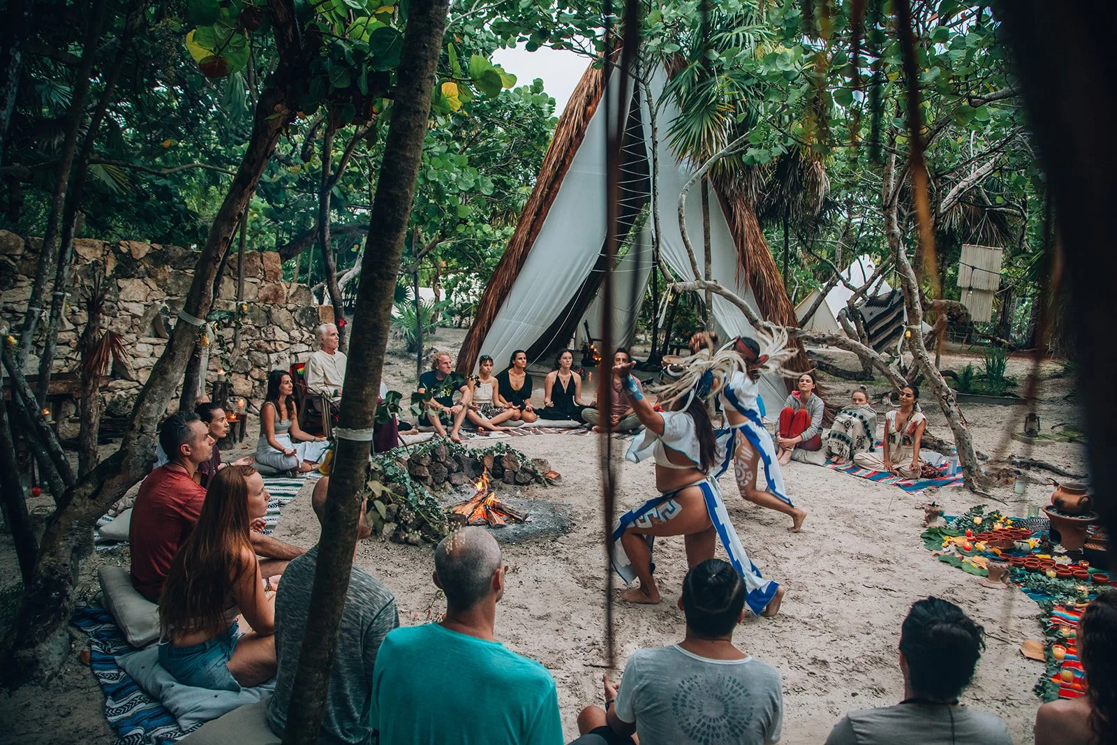 A group of people gathered around a fire in a tropical outdoor setting, watching two women perform a traditional dance wearing feathered headdresses and ceremonial clothing, in front of a teepee-style tent surrounded by lush greenery.