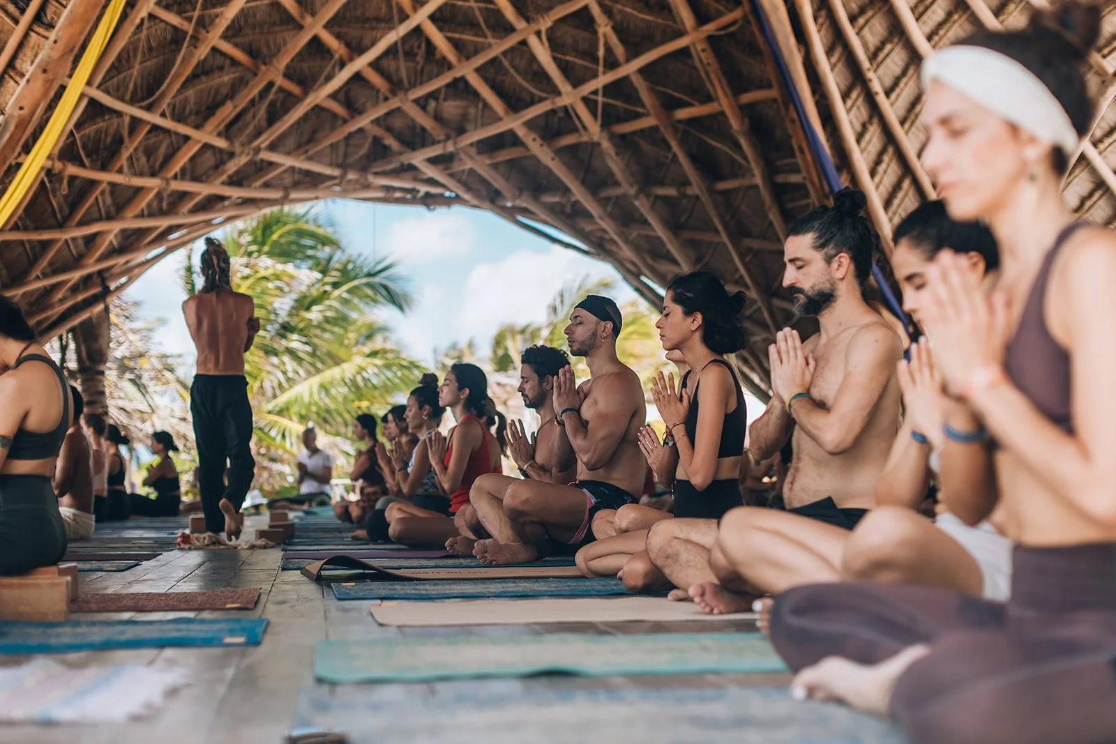 People practicing yoga under a thatched roof with palm trees in the background.