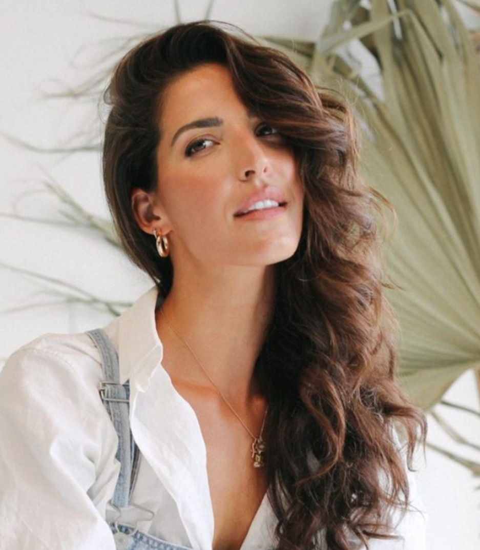 Costa Rica Yoga & Reiki Immersion program. A woman with long, wavy brown hair and hoop earrings smiling slightly at the camera, wearing a white shirt and denim overalls, with large green leaves in the background.