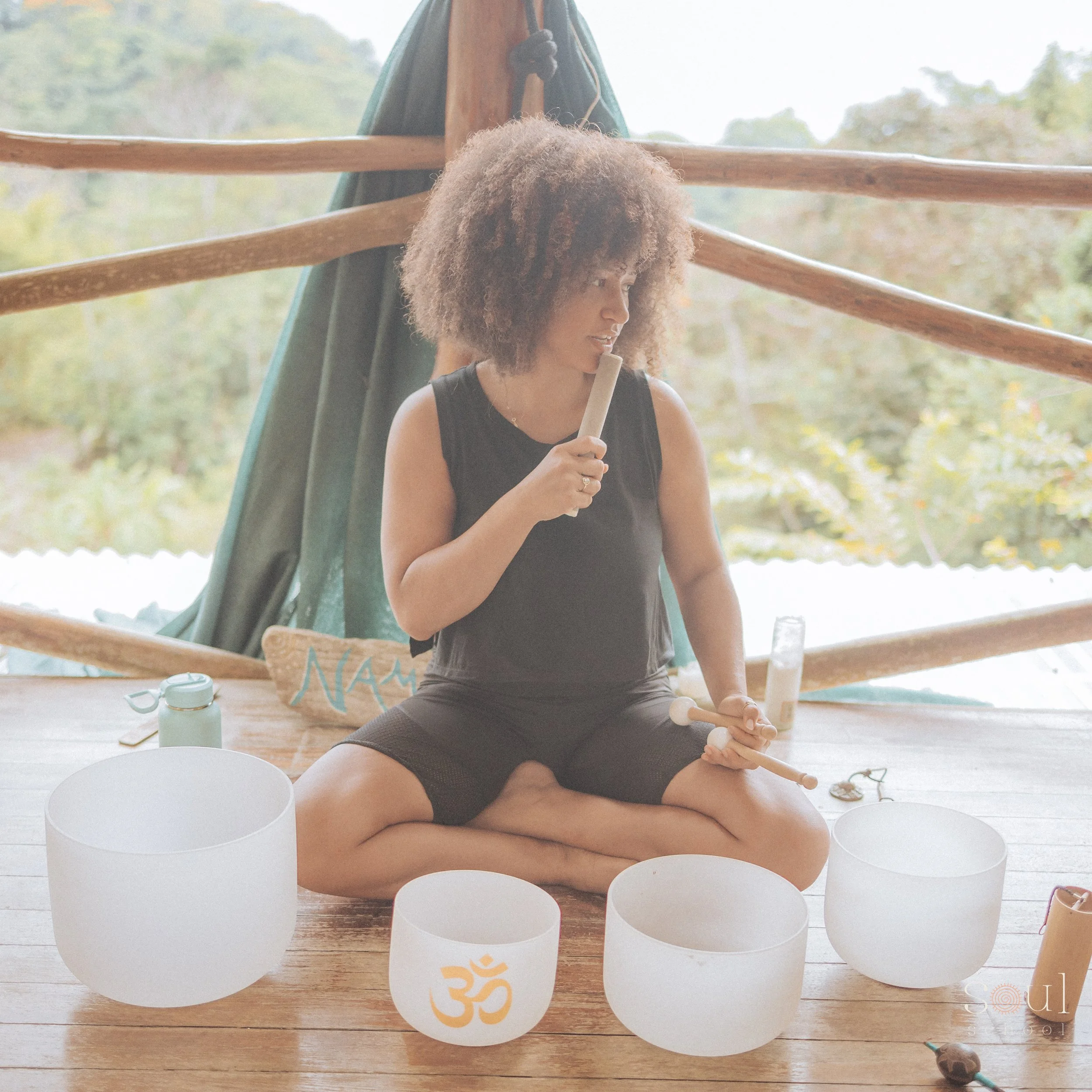 Woman with curly hair sitting cross-legged on wooden floor, playing crystal singing bowls, in an outdoor setting with trees in background.
