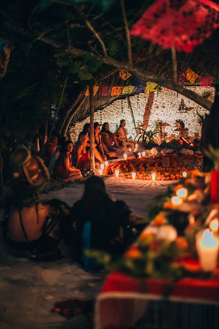 People sitting on the ground in a circle, illuminated by candles, outdoors at night, under trees, with colorful papel picado decorations overhead and a painted brick wall in the background, participating in a cultural or spiritual gathering.