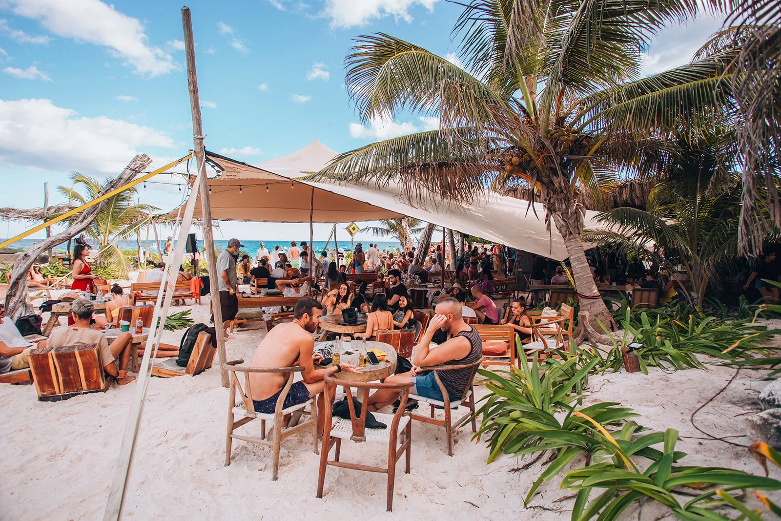 People relaxing and socializing on a sandy beach with palm trees, under large umbrellas, at a beachside bar or restaurant.