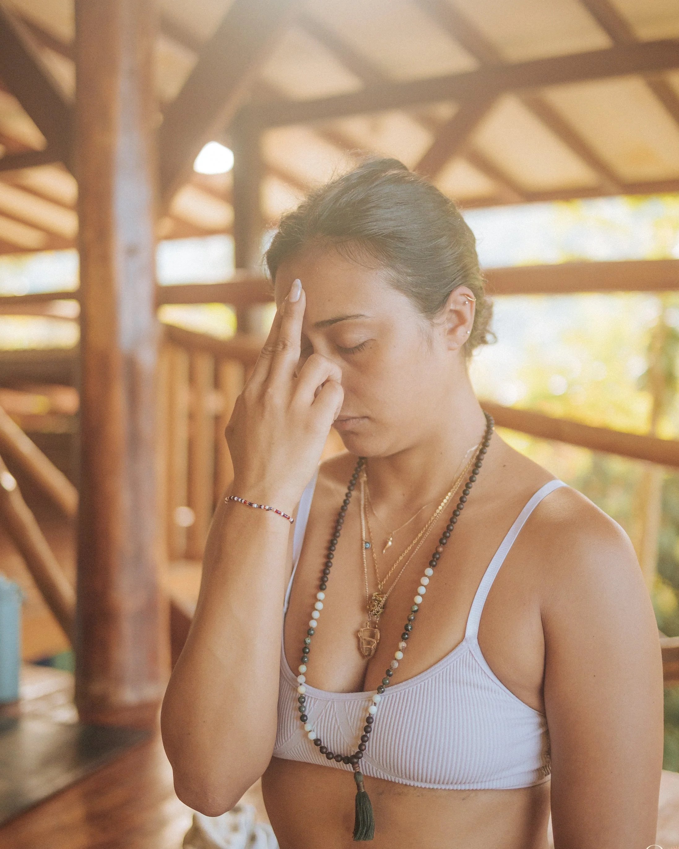 A woman with dark hair tied back, wearing a white tank top and multiple layered necklaces, is indoors with wooden beams in the background. She appears to be meditating or concentrating, with her eyes closed and one hand touching her forehead.