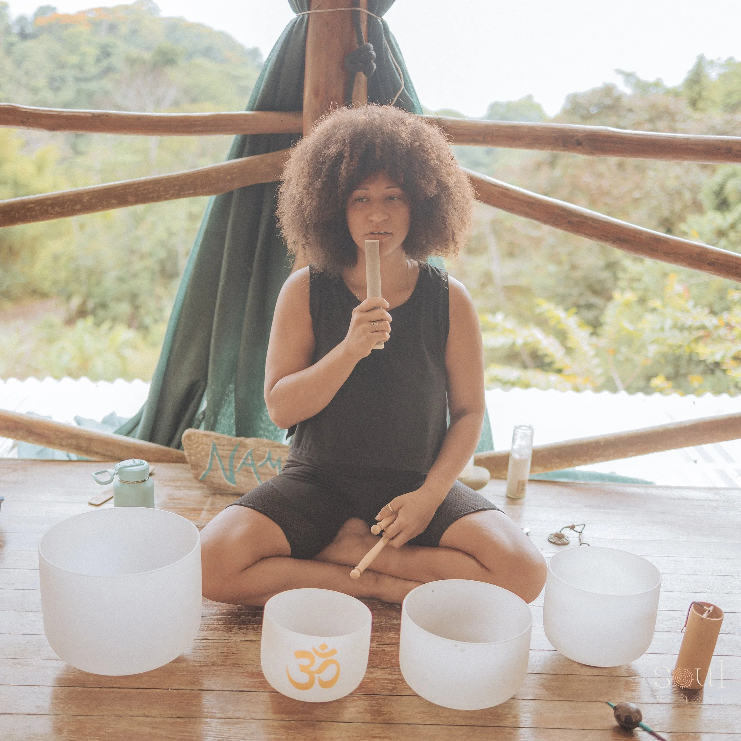 A woman with voluminous curly hair sitting cross-legged on a wooden floor, holding a singing bowl mallet, surrounded by singing bowls, with an outdoor green landscape in the background.