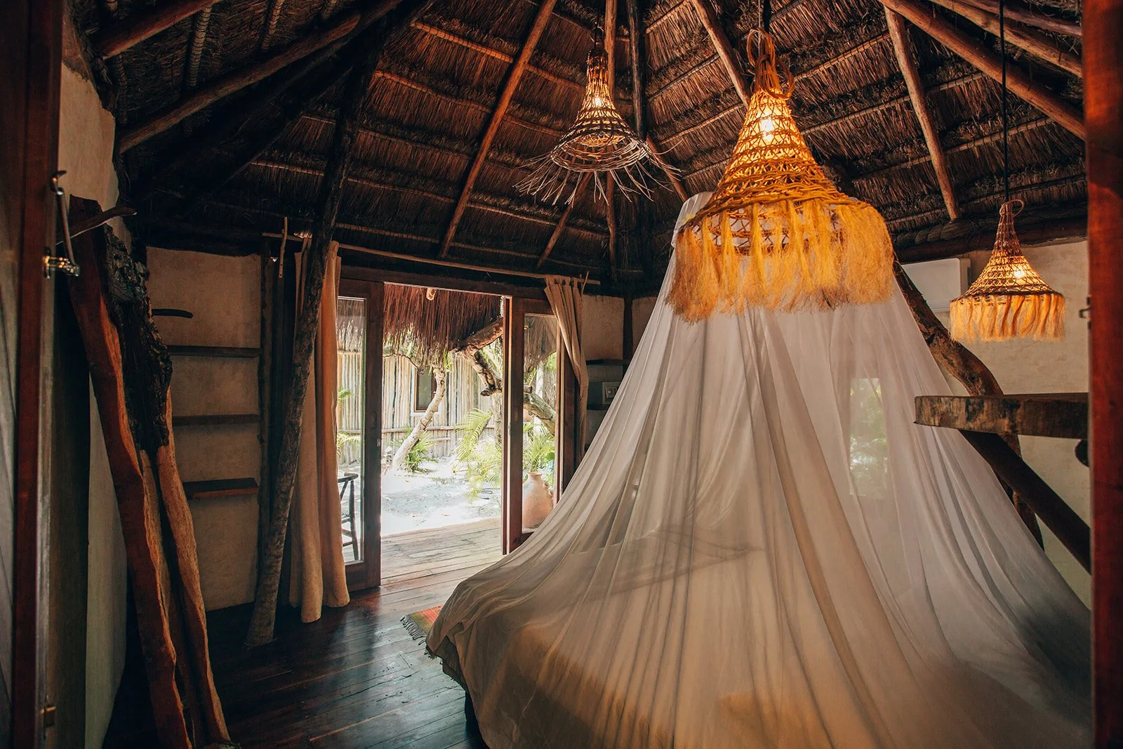 Interior of a rustic wooden hut featuring a bed with a white canopy, wooden ceiling with hanging woven lamps, open sliding door showing a sandy outdoor area with trees and plants.