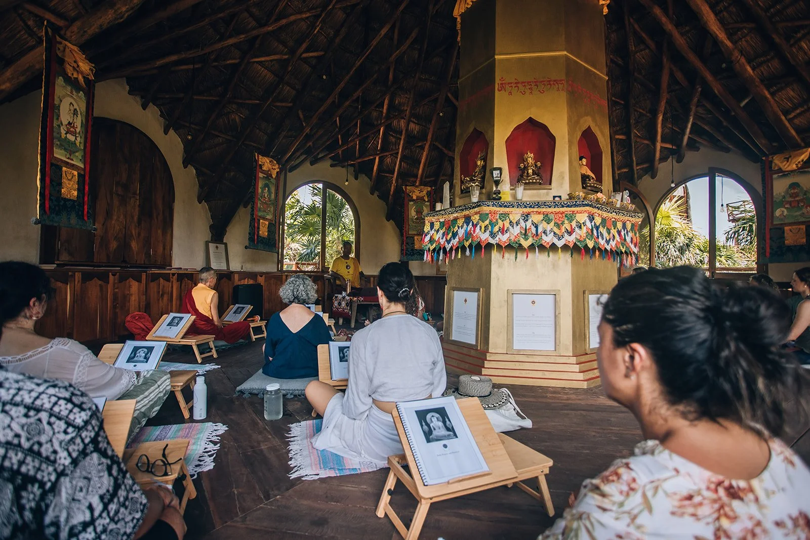 People seated on the floor inside a wooden structure during a meditation or spiritual ceremony, with a central altar with statues and colorful decorations, and open windows showing greenery outside.