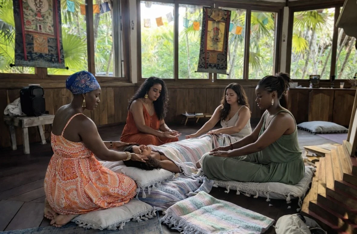 Four women of diverse ethnicities participating in a spiritual or meditation ritual inside a wooden, tropical-style room with large windows and green foliage outside. One woman is lying down with her eyes closed, while the others sit around her with their hands extended towards her, perhaps performing a healing or blessing.