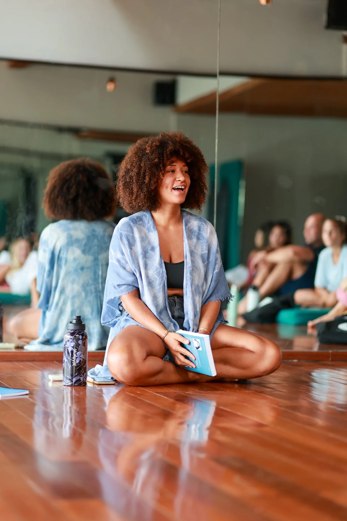 Young woman sitting cross-legged on wooden floor, holding a book, smiling during a group activity in a mirrored room.